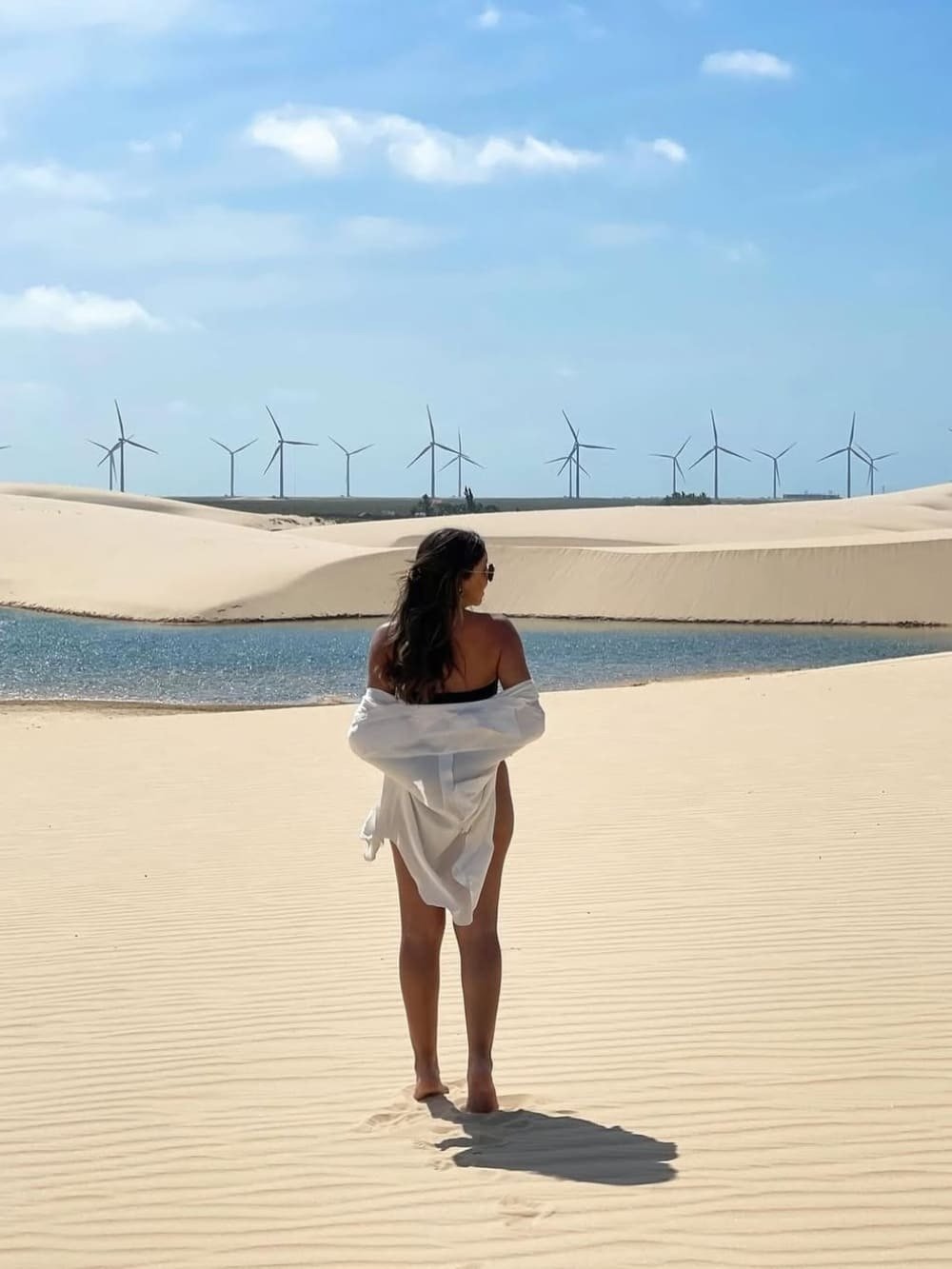 Sand, water, wind — the dunes hold it all at once. A woman in a white shirt stands barefoot on rippled white sand dunes facing a blue lagoon, with a row of wind turbines on the horizon — a striking scene from the Lençóis Maranhenses full itinerary