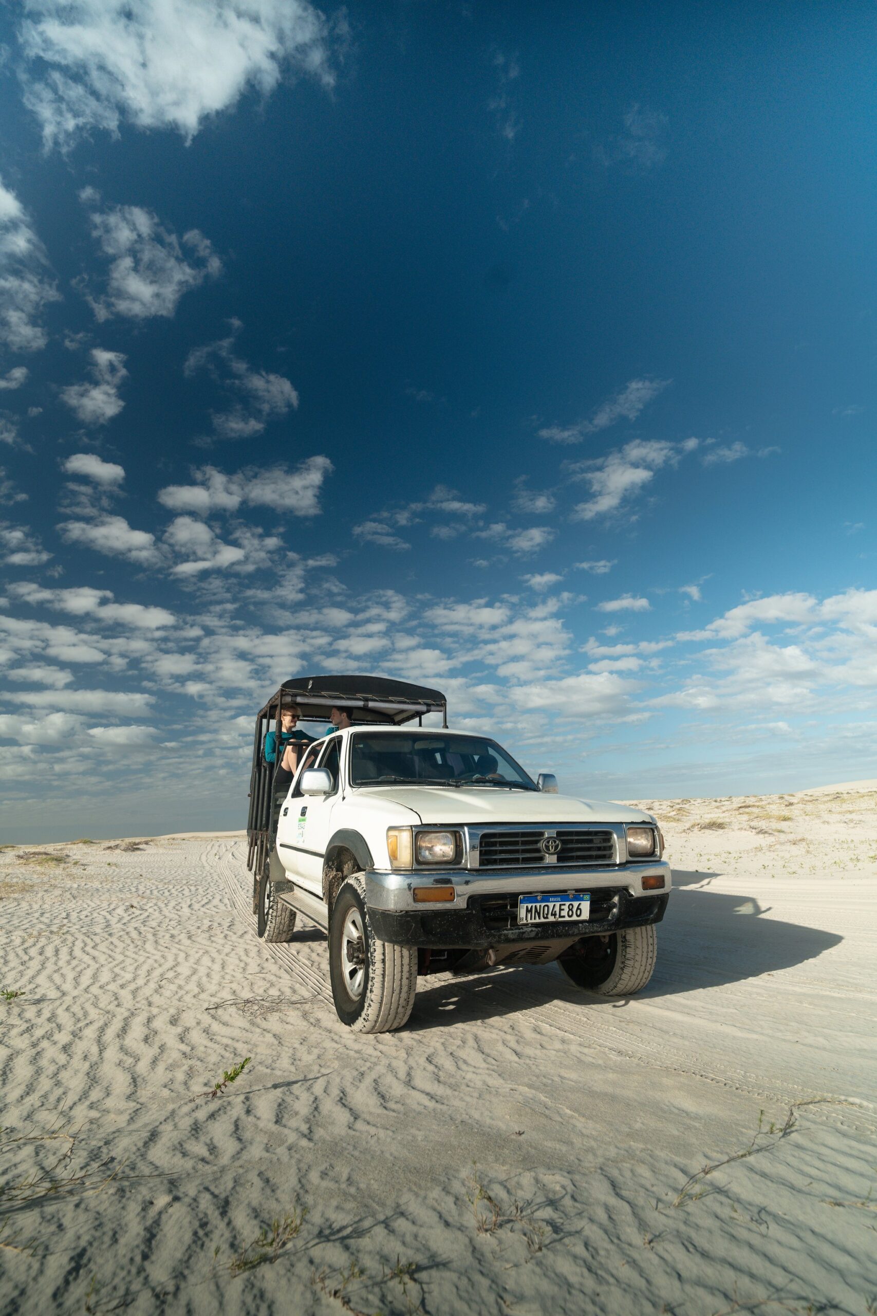 Carroceria aberta, areia ondulada e duinas sem fim à frente. Uma picape 4x4 branca com carroceria aberta transporta passageiros pelas vastas dunas de areia branca dos Lençóis Maranhenses sob um céu azul dramático com nuvens esparsas