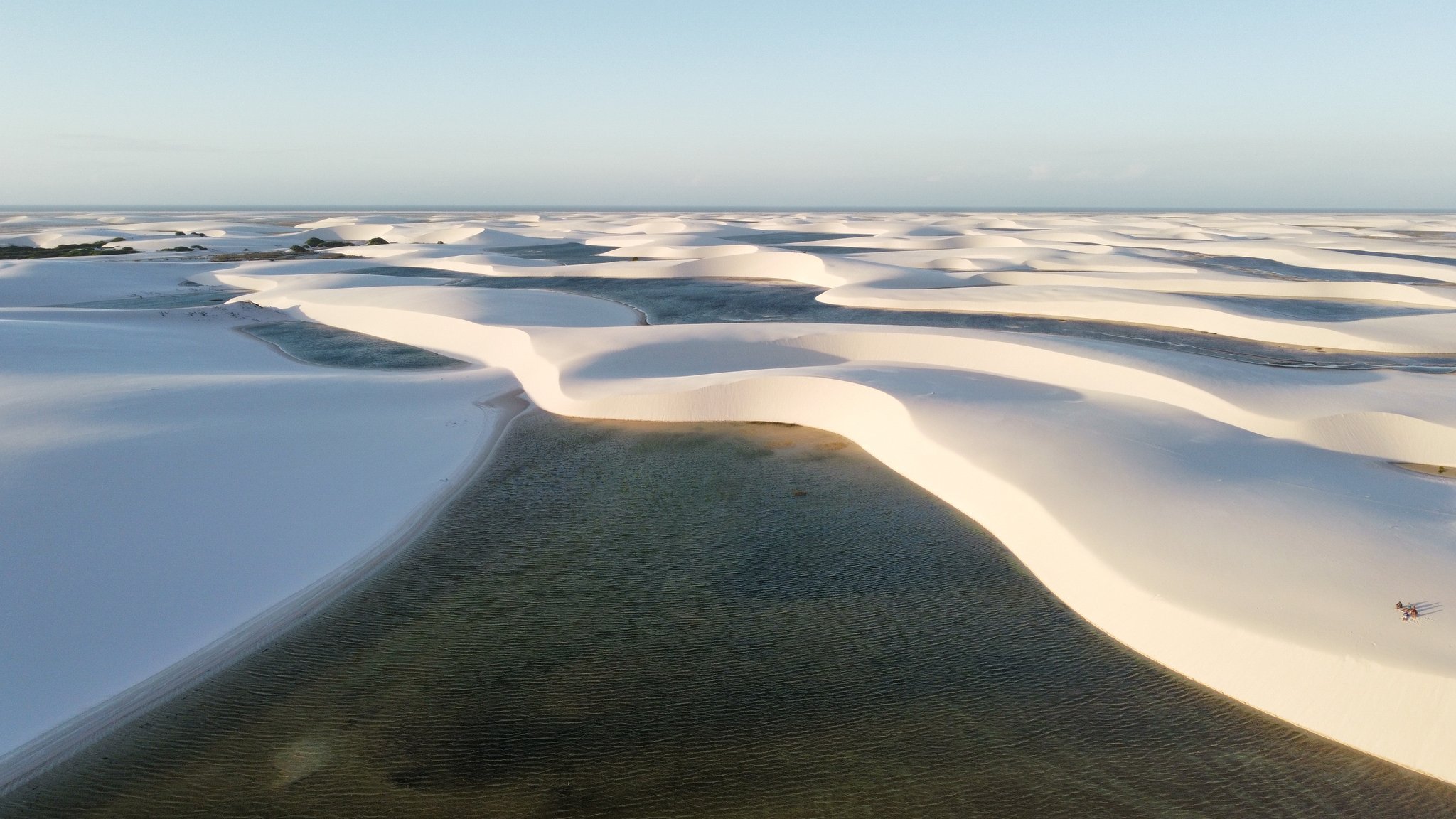 Dunas sem fim, uma lagoa verde espelhada — o parque visto de cima revela sua grandiosidade. Vista aérea da trilha pelos Lençóis Maranhenses, com vastas dunas de areia branca contornando uma lagoa de água doce verde-escura que se estende até o horizonte sob um céu azul claro