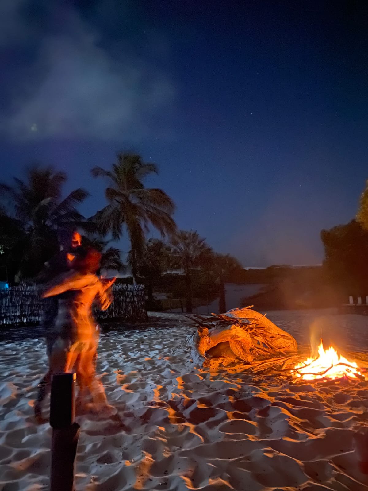 Chamas, areia branca e um céu repleto de estrelas — a noite é toda sua. Uma figura em movimento ao lado de uma fogueira brilhante em uma praia de areia branca à noite, com palmeiras em silhueta contra um céu azul profundo estrelado na trilha pelos Lençóis Maranhenses