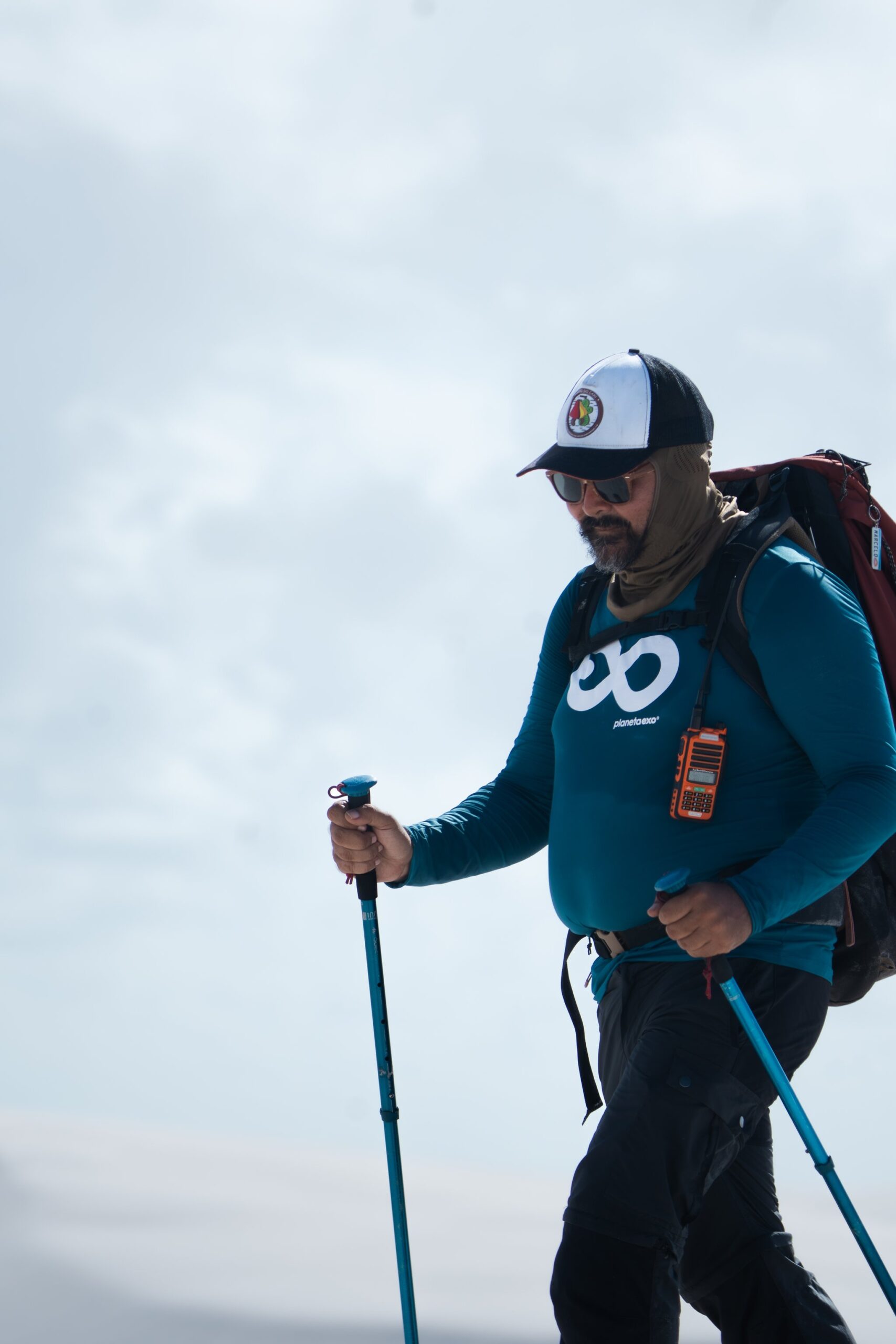 Bastões no chão, rádio no peito, mochila pesada — o guia abre o caminho. Um guia da PlanetaEXO com bastões de caminhada, mochila grande e rádio lidera a trilha nos Lençóis Maranhenses sob céu nublado