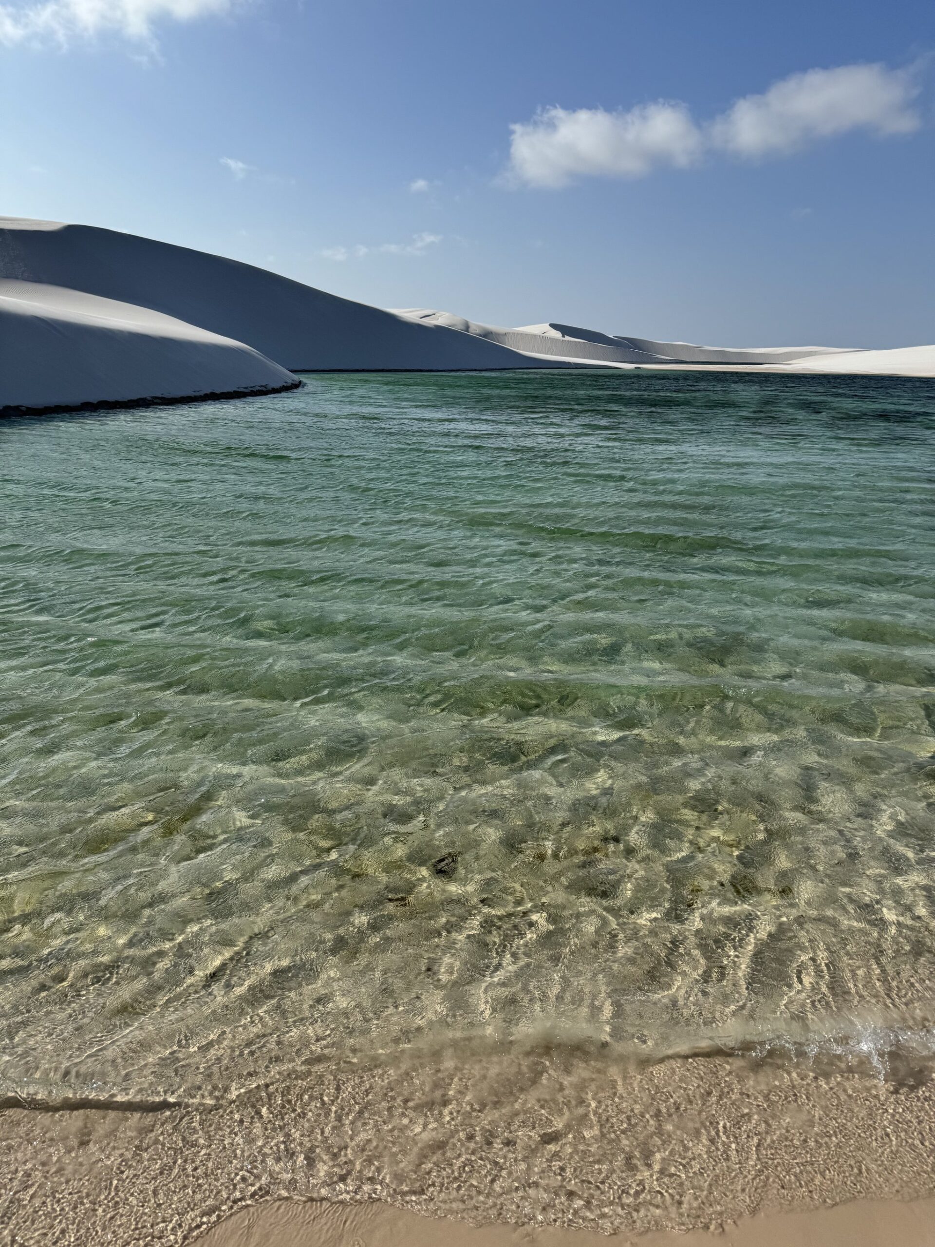 Água transparente, dunas brancas ao fundo — a recompensa perfeita depois de uma longa caminhada. Lagoa de água doce turquesa e cristalina banhando a areia durante uma trilha nos Lençóis Maranhenses, com dunas de areia branca se erguendo sob um céu azul ao fundo