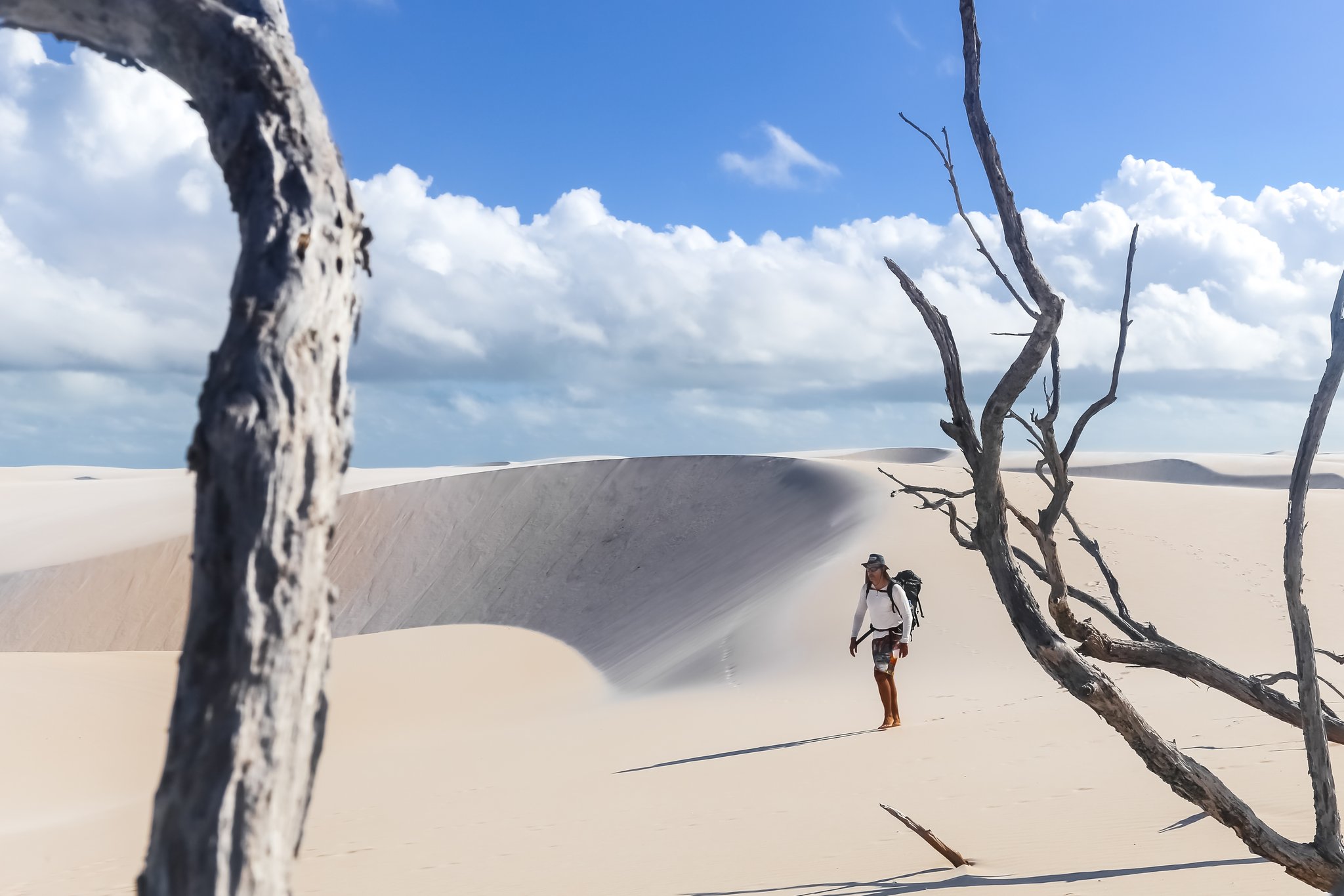 Galhos secos, areia branca, um caminhante engolido pelas dunas. Photo: MaRabelo Um mochileiro de chapéu faz uma pausa entre árvores mortas e branqueadas sobre uma vasta duna de areia branca durante uma trilha nos Lençóis Maranhenses, com cristas de dunas esculpidas sob um céu azul intenso