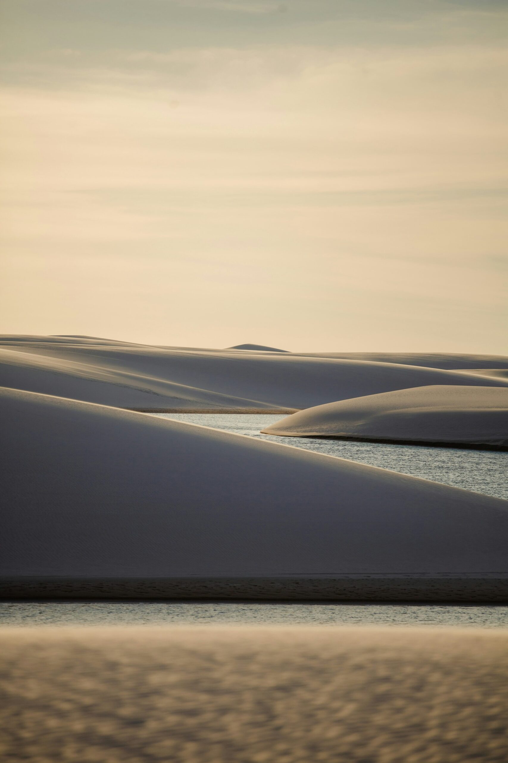 Silêncio em camadas: dunas brancas, água parada, céu que desbota. Dunas de areia branca lançando sombras azuis profundas sobre lagoas tranquilas na luz suave do entardecer nos Lençóis Maranhenses