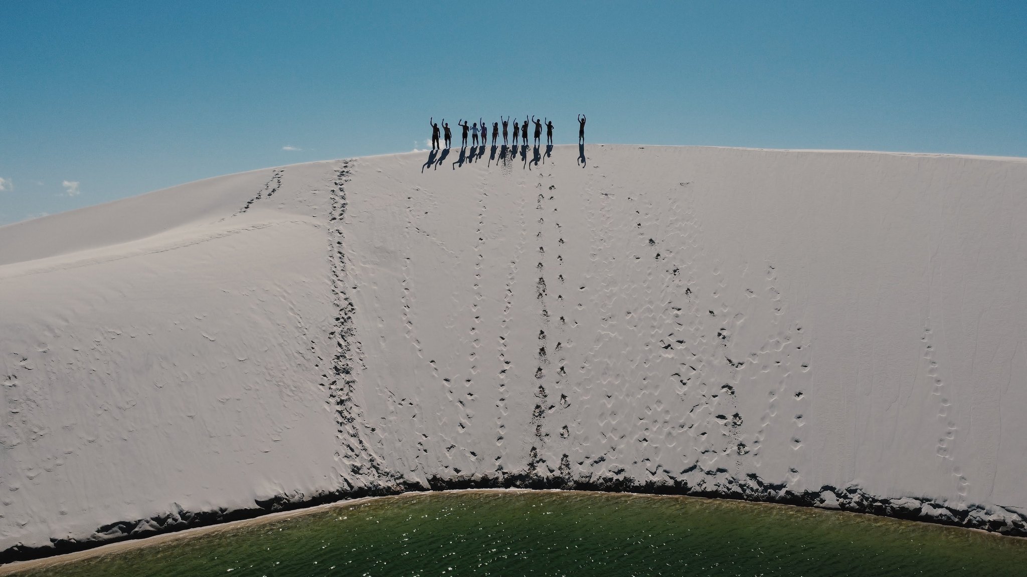 Doze silhuetas no alto da duna, braços erguidos, lagoa verde lá embaixo. Um grupo de caminhantes em silhueta contra um céu azul no topo de uma enorme duna de areia branca durante uma trilha nos Lençóis Maranhenses, com uma lagoa verde visível ao pé da duna
