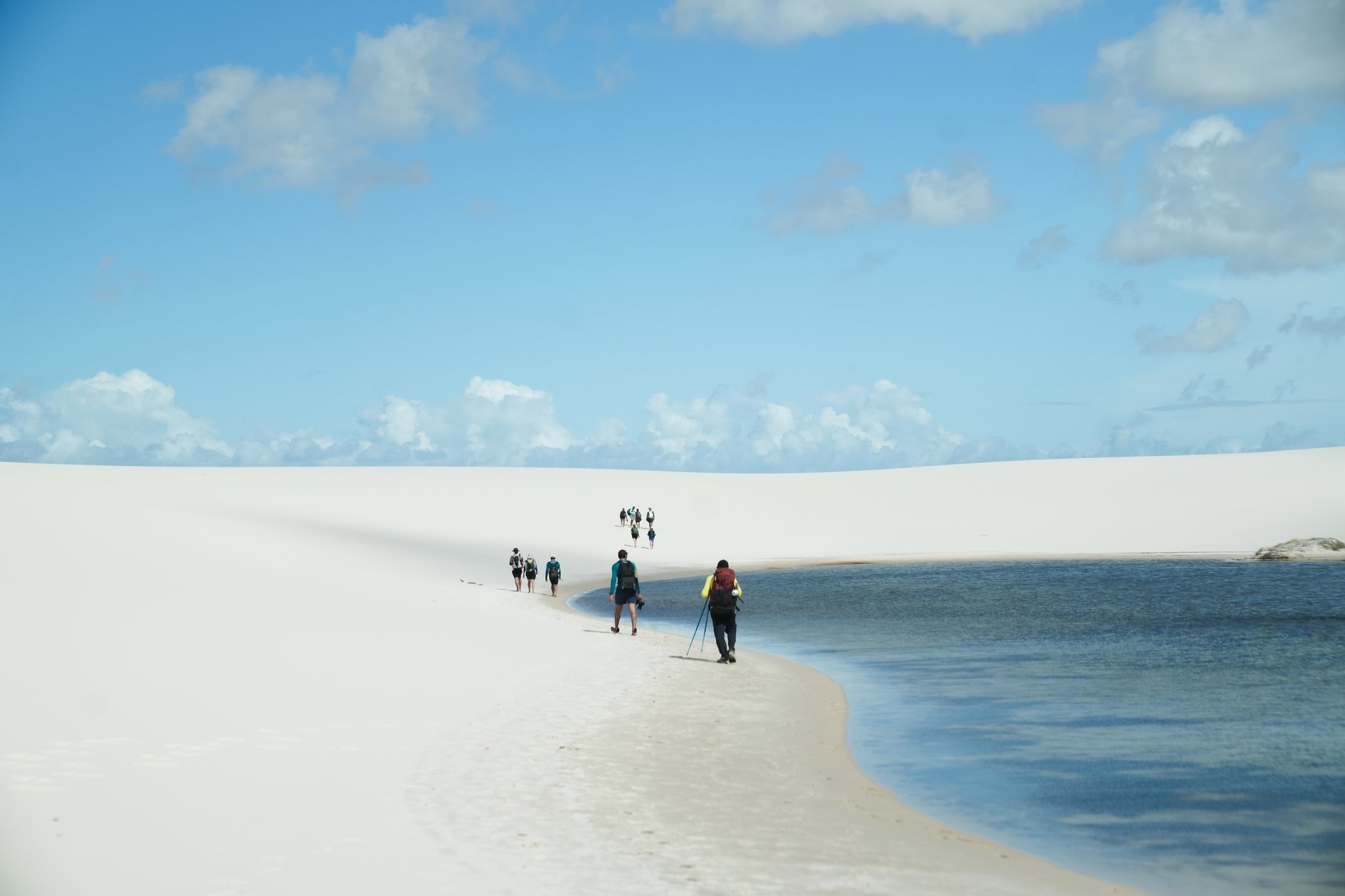 Um grupo de caminhantes com mochilas avança em fila indiana por uma estreita faixa de areia branca às margens de uma lagoa de água doce durante uma trilha nos Lençóis Maranhenses, com dunas e céu azul ao fundo