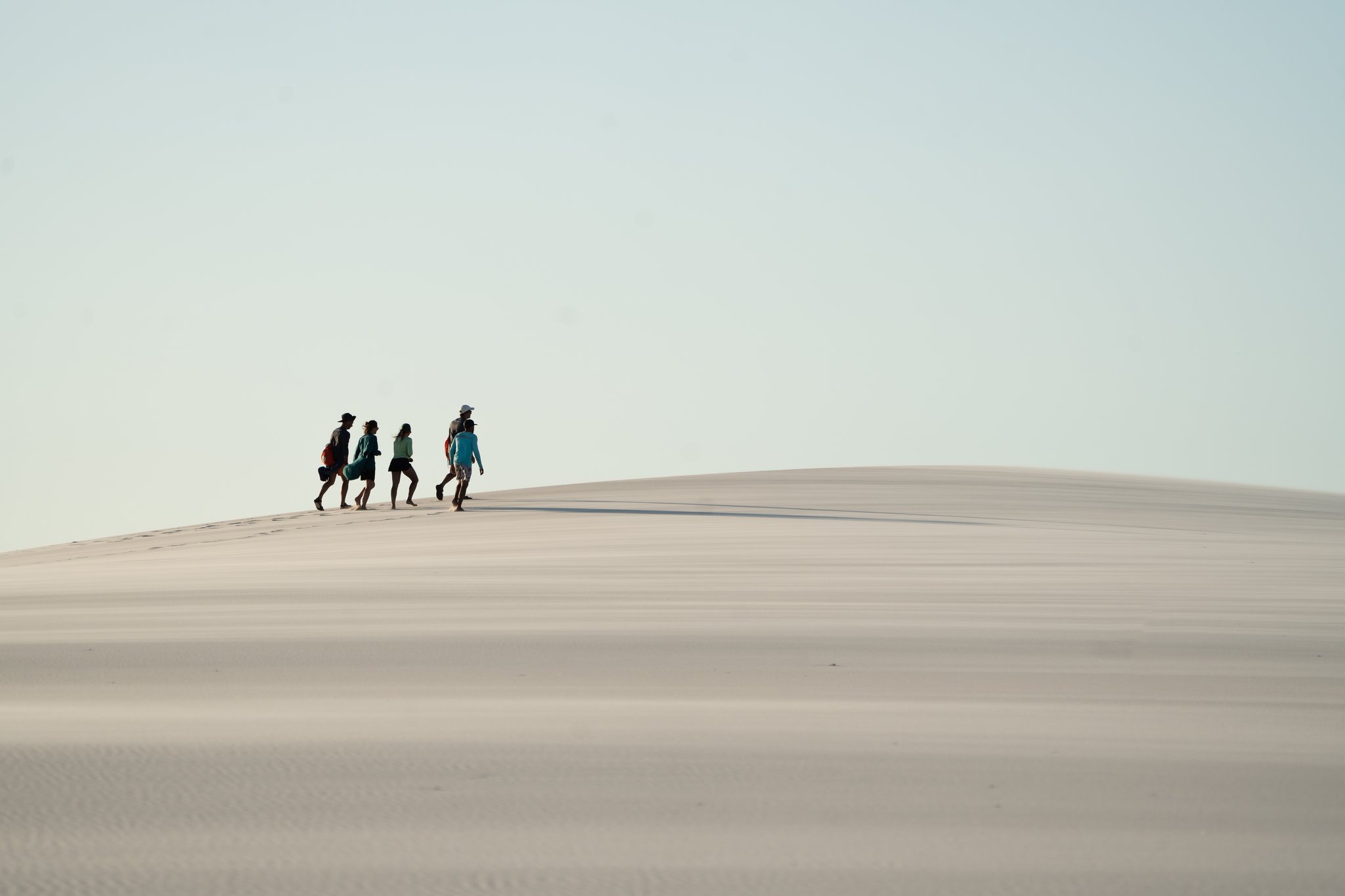 Quatro figuras, uma duna sem fim — a imensidão dos Lençóis resumida numa só cena. Quatro caminhantes percorrem o cume de uma imensa duna de areia branca durante uma trilha nos Lençóis Maranhenses, figuras diminutas contra um céu aberto e claro