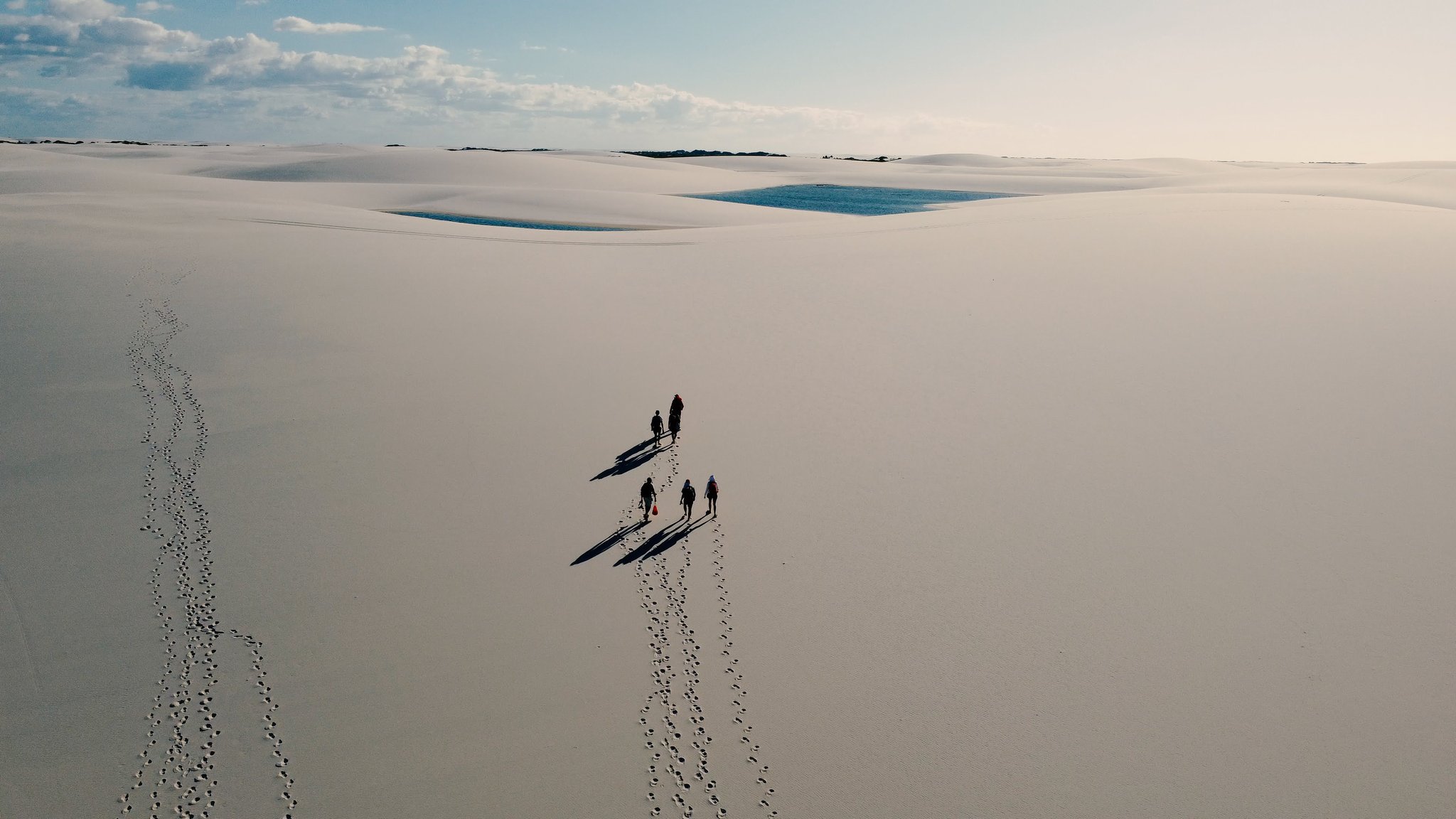 Cinco figuras, areia branca sem fim e duas lagoas chamando à frente. Vista aérea de cinco caminhantes projetando longas sombras sobre as dunas brancas dos Lençóis Maranhenses, com duas lagoas turquesa reluzindo ao fundo