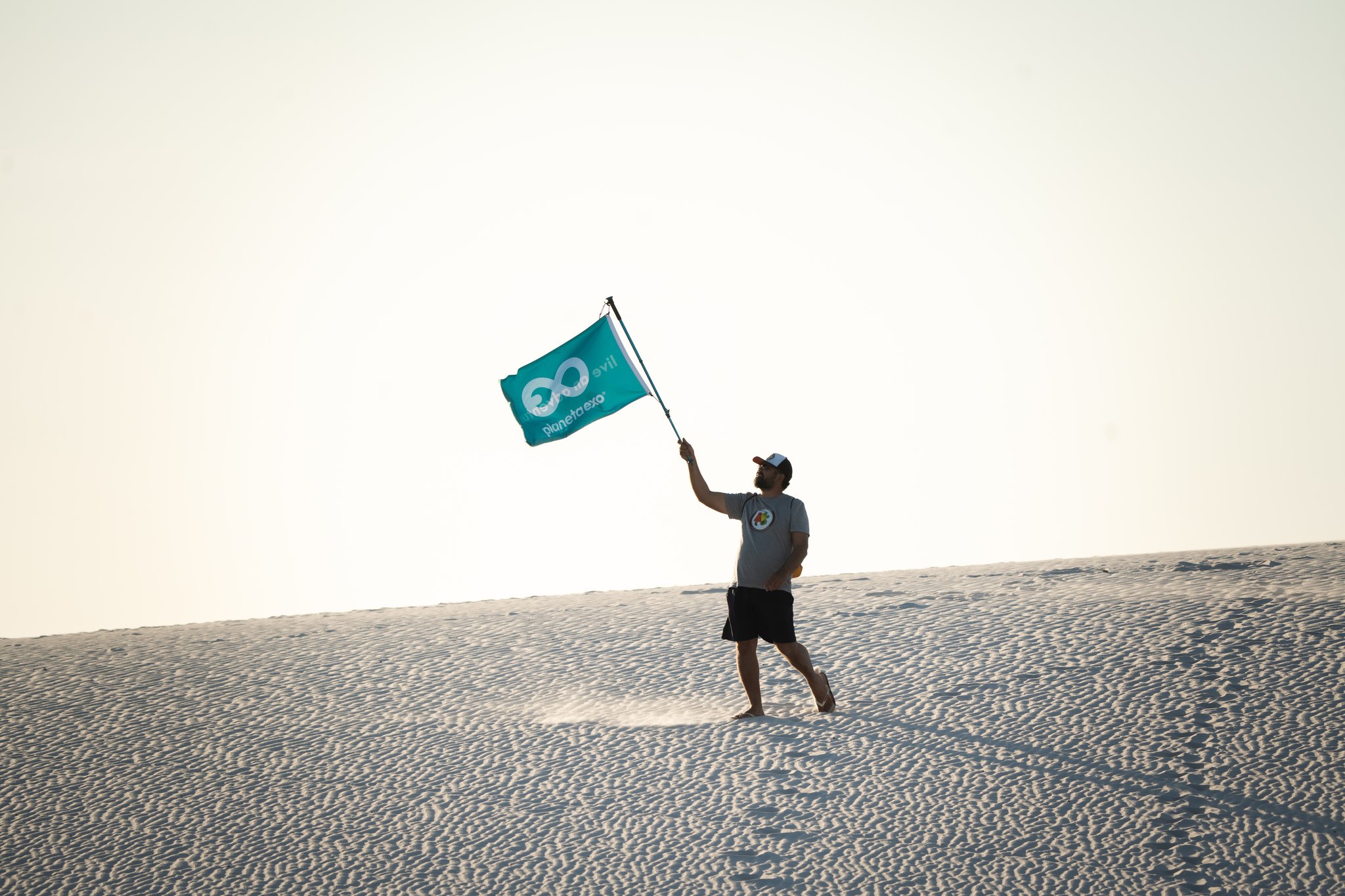 Um guia agita uma bandeira turquesa da PlanetaEXO caminhando pelas imensas dunas brancas dos Lençóis Maranhenses