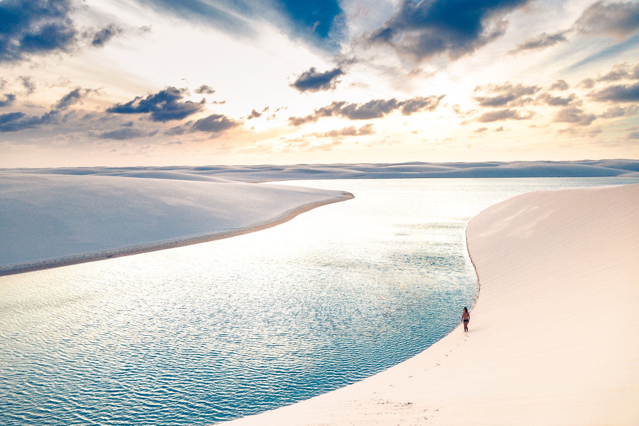 Uma figura pequena, dunas imensas e a luz dourada do fim do dia. Photo: cokada Um caminhante solitário percorre a borda curva de uma lagoa de água doce cristalina entre dunas brancas na trilha pelos Lençóis Maranhenses, sob um céu dourado ao entardecer