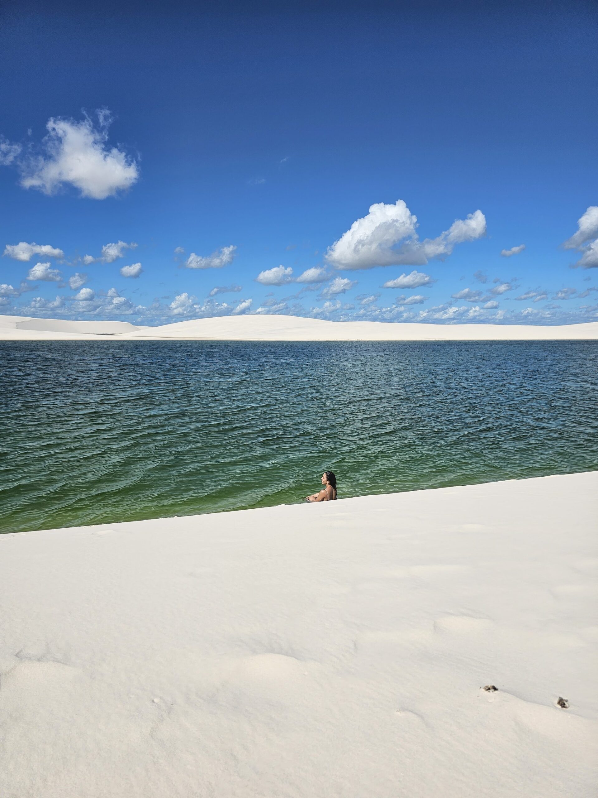 Mergulhada no verde, rodeada pelo branco infinito das dunas. Uma nadadora solitária até a cintura em uma ampla lagoa de água doce verde na travessia dos Lençóis Maranhenses, emoldurada por dunas brancas e um céu azul intenso