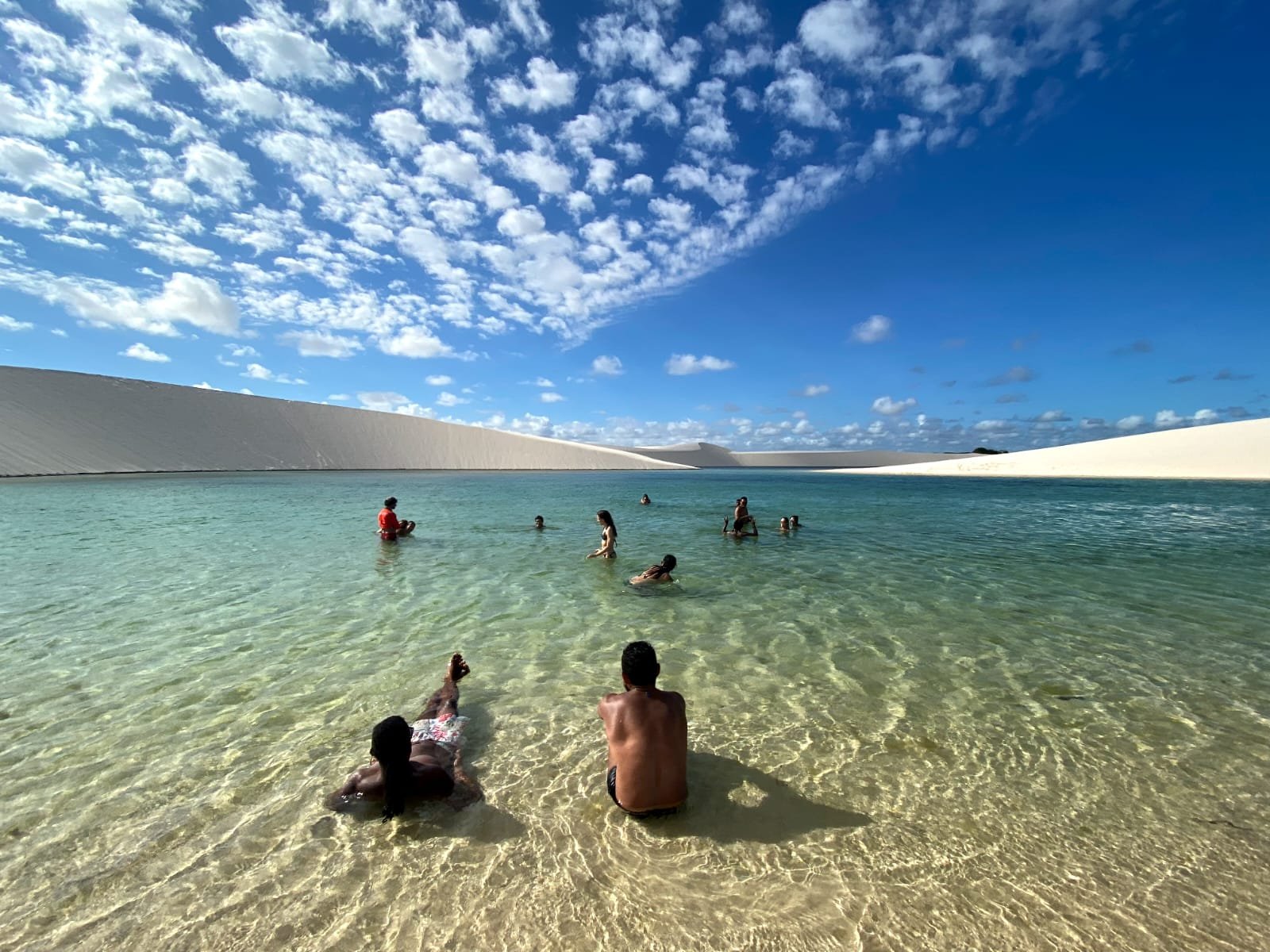 Mergulho merecido após quilômetros de areia branca. Um grupo de viajantes mergulha e relaxa em uma lagoa de água doce cristalina durante uma trilha pelos Lençóis Maranhenses, cercados por dunas de areia branca sob um céu azul intenso