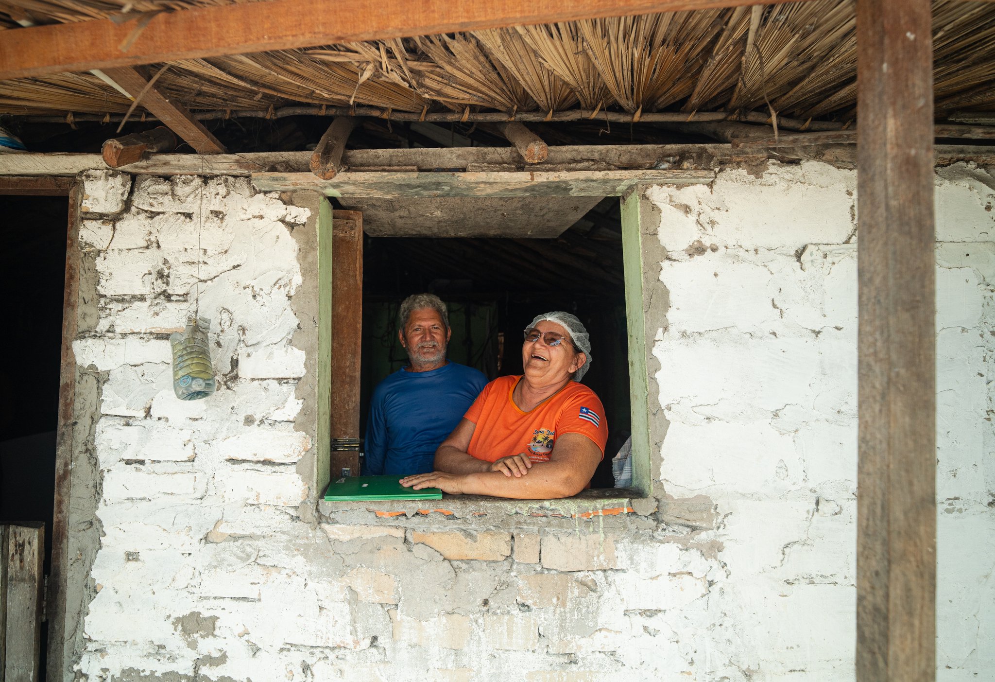 Risadas à janela — a acolhida das famílias dos oásis é o coração desta aventura. Um homem sorridente e uma mulher gargalhando se apoiam na janela de uma casa de tijolos caiados sob telhado de palha, recebendo com alegria os visitantes durante a travessia dos Lençóis Maranhenses
