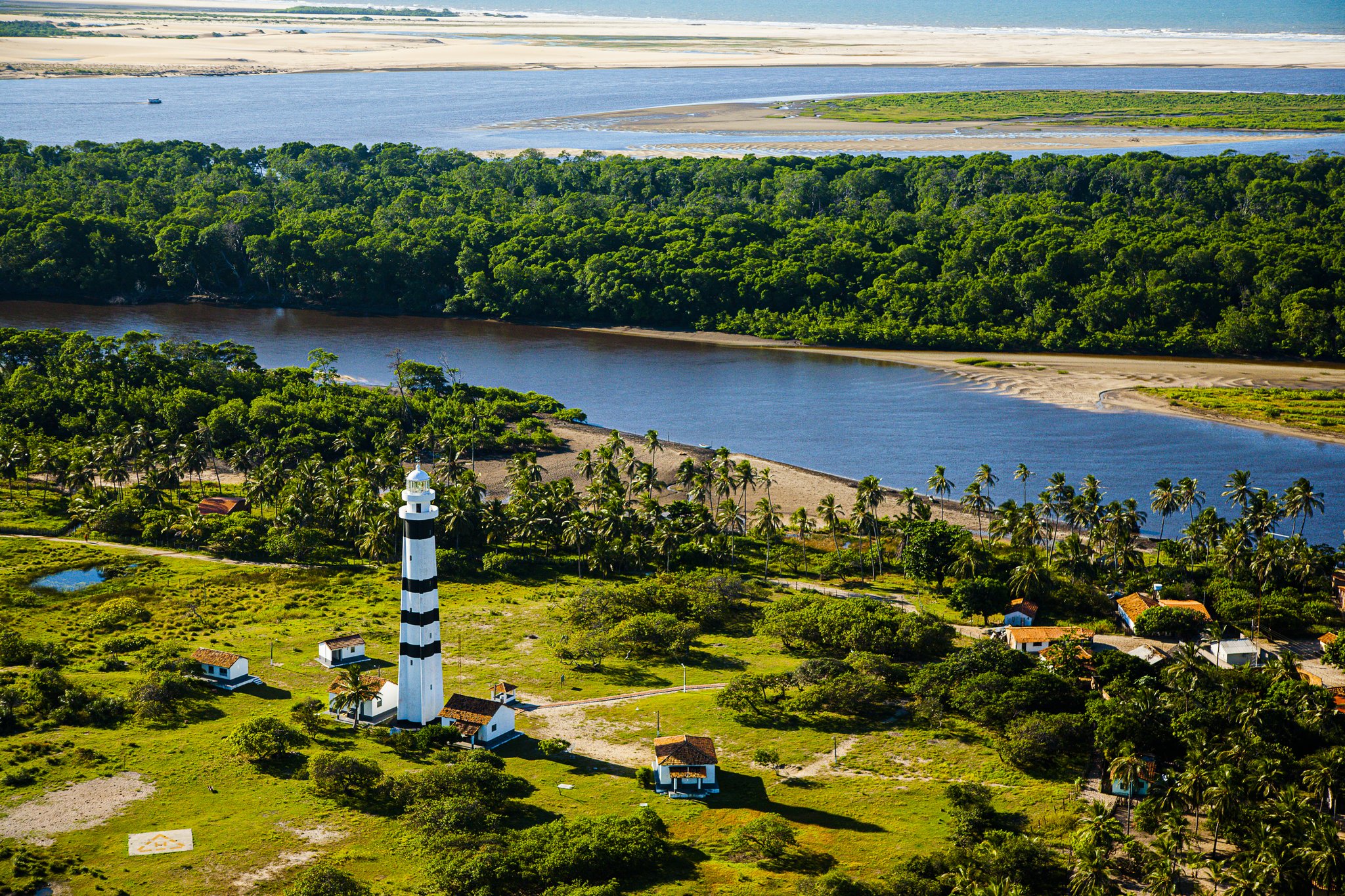 Farol listrado, coqueiros e dunas num só panorama. Photo: Gabriel Boieras Vista aérea de um farol listrado em preto e branco cercado por palmeiras e vegetação densa, com um amplo estuário fluvial e dunas de areia branca se estendendo até o horizonte — marco da rota de trilha nos Lençóis Maranhenses