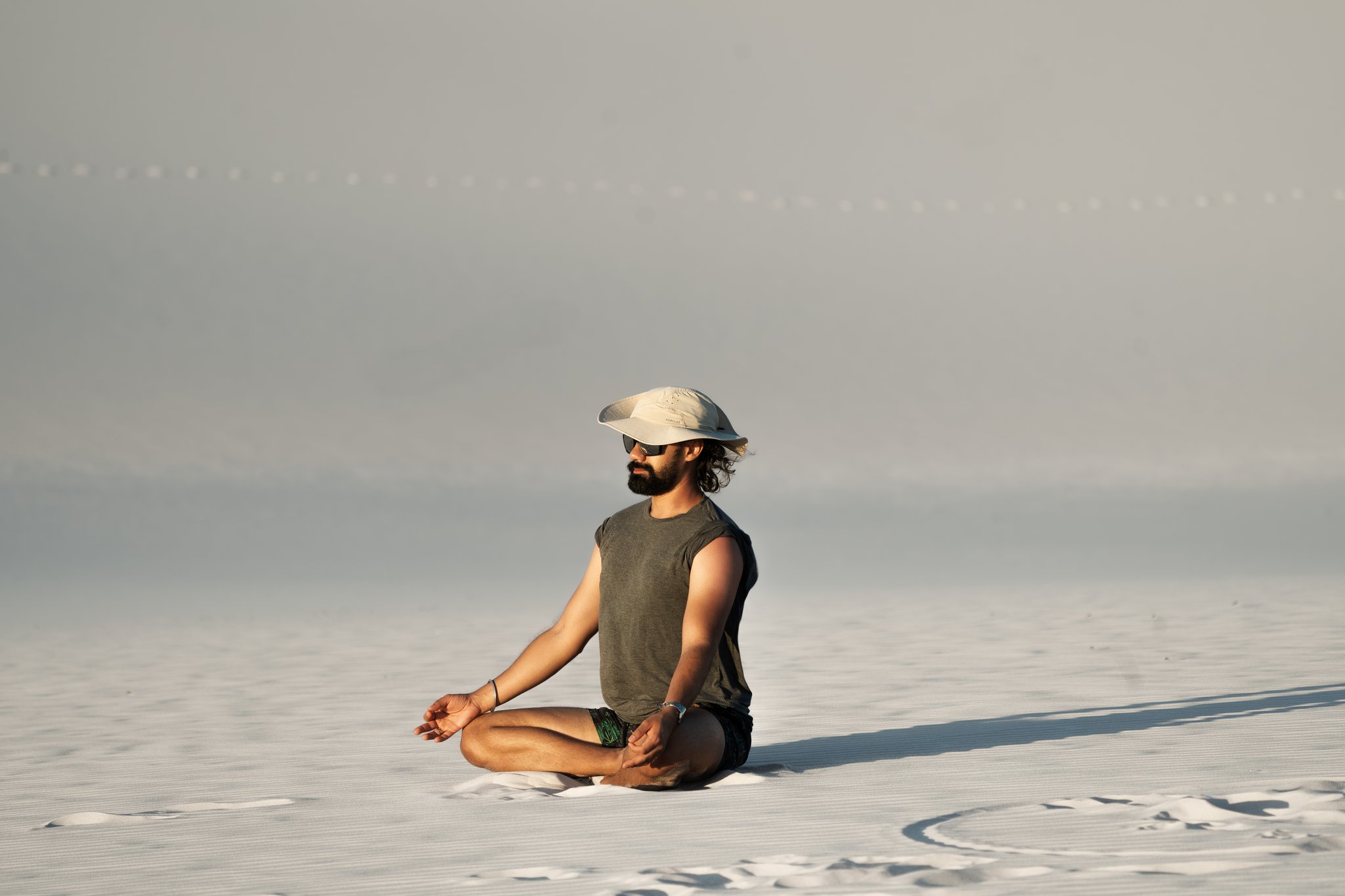 Silêncio de corpo e mente no coração dos lençóis brancos. Um homem barbudo com boné e óculos de sol senta-se de pernas cruzadas sobre o imenso tapete branco de areia durante a travessia a pé dos Lençóis Maranhenses, envolto por um horizonte pálido e infinito