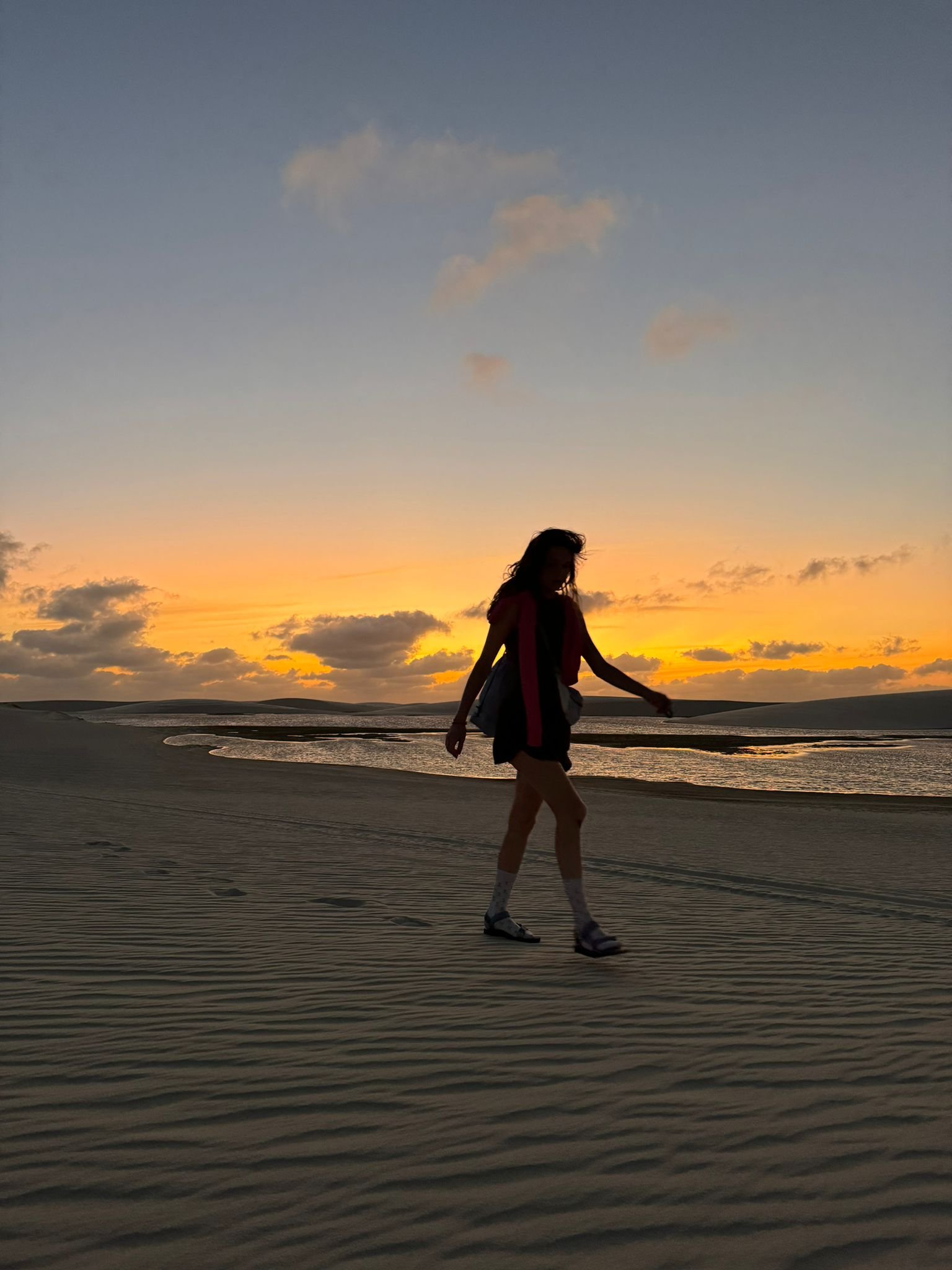 Areia rendada pelo vento, céu em brasa — o fim de tarde que vale cada passo. Silhueta de uma mulher caminhando sobre dunas de areia ondulada pelo vento durante uma trilha nos Lençóis Maranhenses, com um pôr do sol alaranjado e uma lagoa tranquila ao fundo