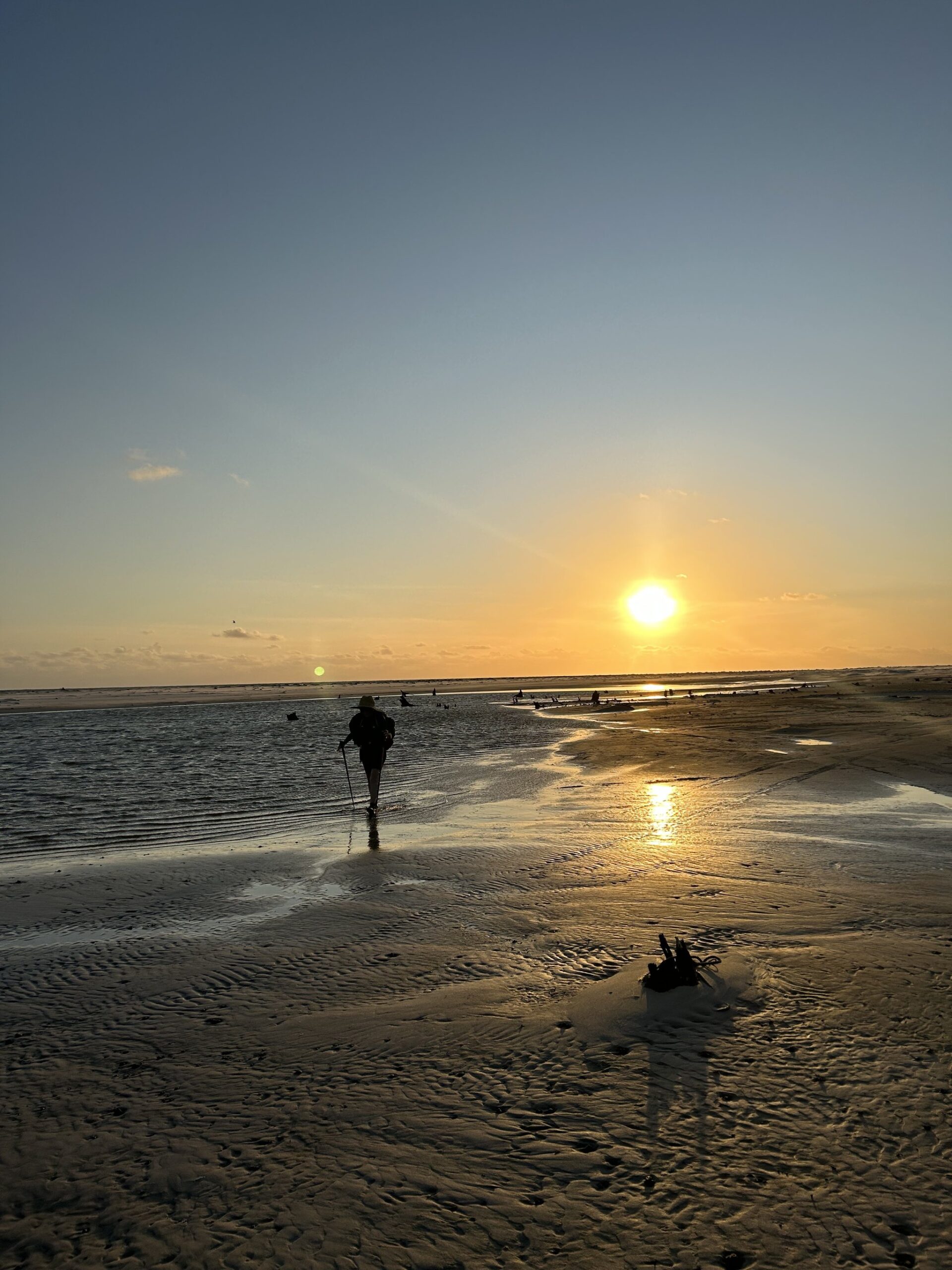 Areia molhada, céu em brasa — o fim do dia que recompensa cada passo. Um caminhante solitário com mochila e bastões de trekking atravessa águas rasas na trilha pelos Lençóis Maranhenses, em silhueta contra um pôr do sol dourado sobre extensas planícies de areia ripada