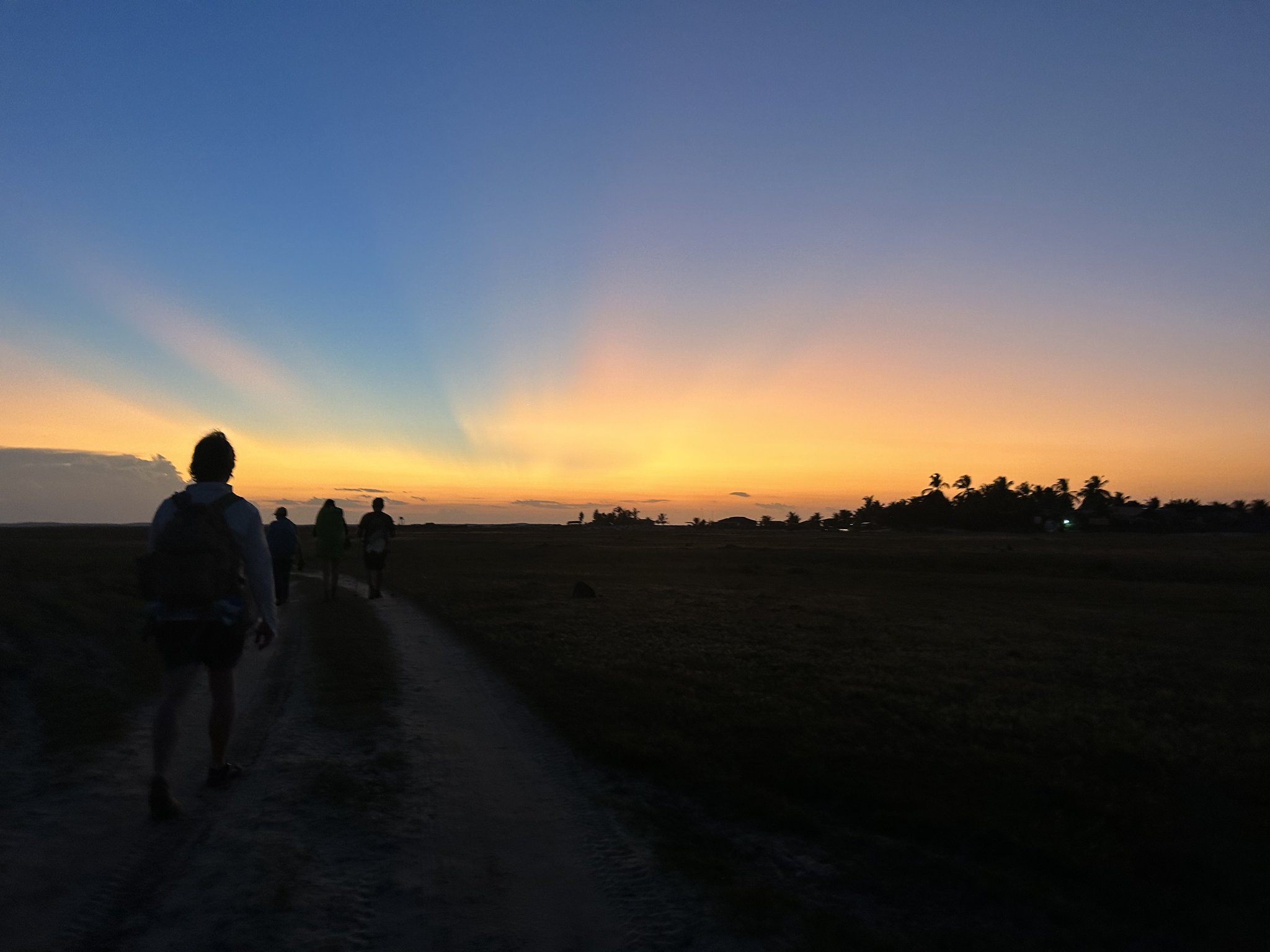 A planície se apaga no entardecer e a trilha segue adiante. Silhuetas de caminhantes em uma trilha de areia durante a travessia dos Lençóis Maranhenses ao pôr do sol, com tons laranja e azul e palmeiras no horizonte
