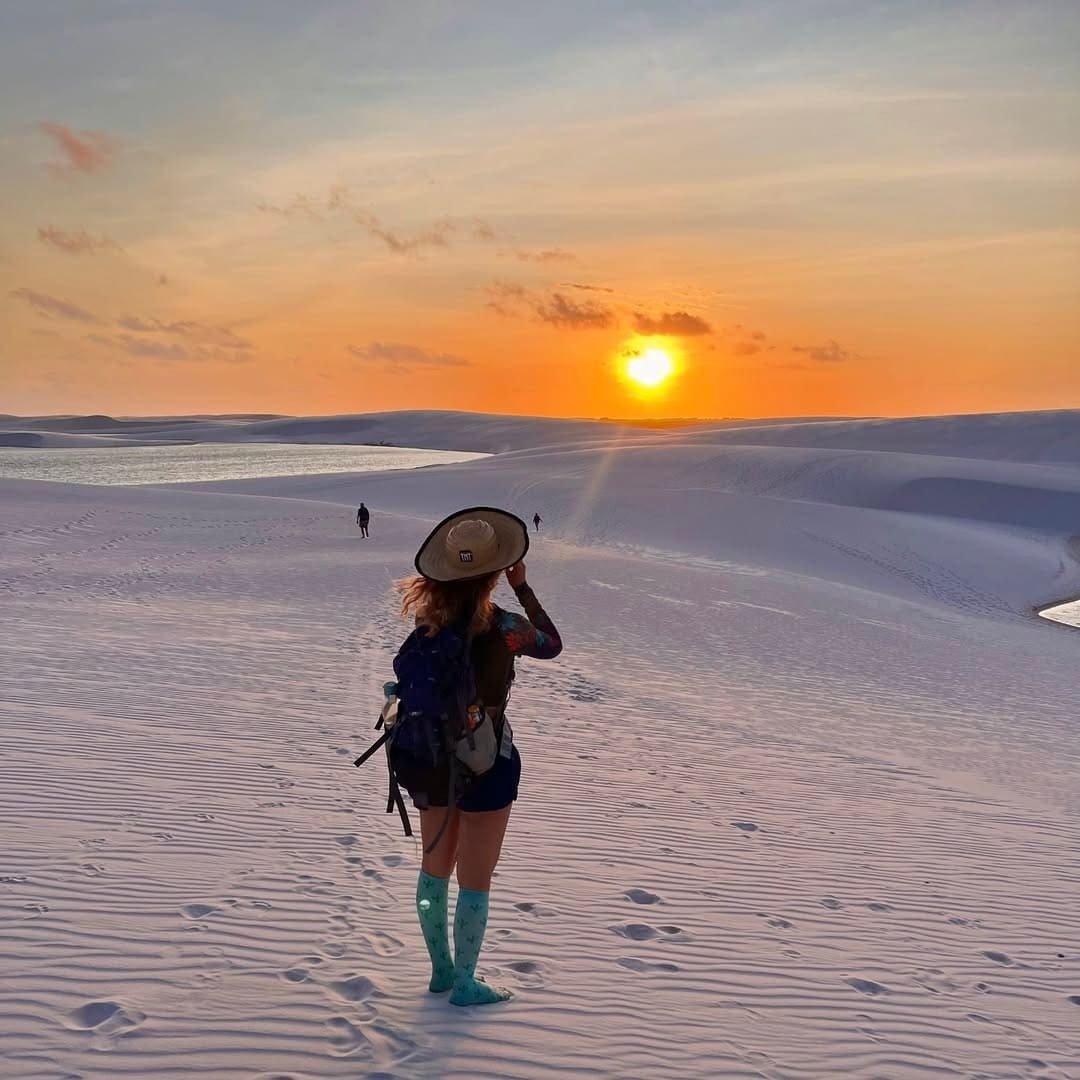 Chapéu firme contra o vento, o horizonte todo em brasa. Uma caminhante com mochila e chapéu de abas largas parada sobre as dunas brancas durante a trilha nos Lençóis Maranhenses, contemplando um pôr do sol alaranjado com uma lagoa de água doce brilhando ao fundo