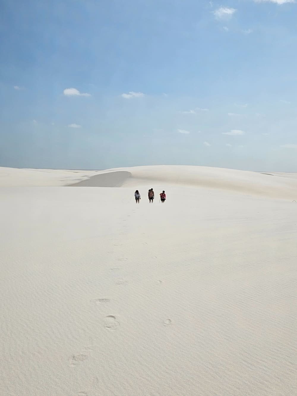 Pegadas no branco infinito, três caminhantes se perdem entre as dunas. Três caminhantes atravessam imensas dunas brancas na trilha pelos Lençóis Maranhenses, com suas pegadas marcadas na areia sob um céu azul claro