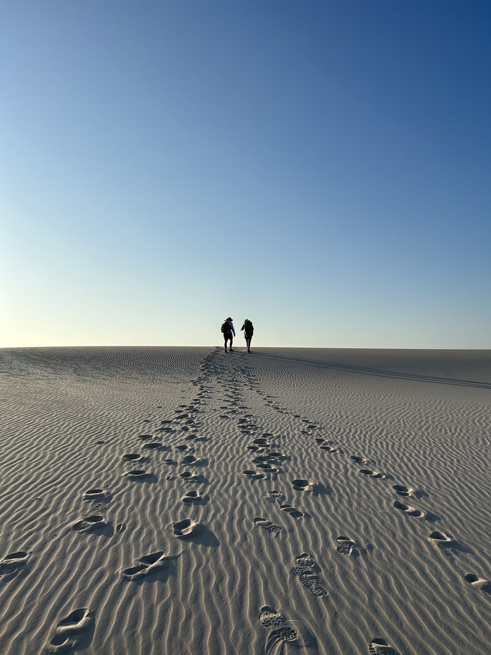 Areia branca, pegadas e o horizonte chamando. Duas silhuetas de caminhantes com mochilas caminham em direção ao horizonte sobre uma vasta duna de areia branca e ondulada durante uma trilha nos Lençóis Maranhenses, com pegadas se estendendo pelo primeiro plano sob um céu azul límpido