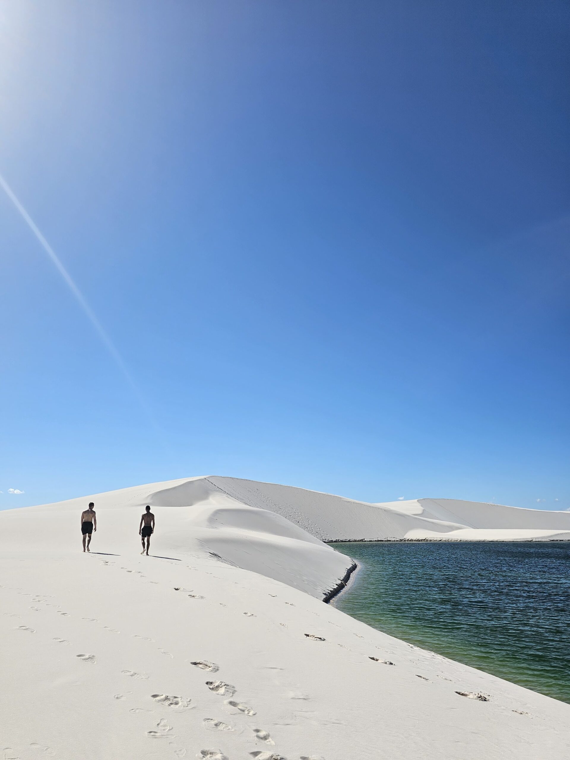 Pegadas à frente, água fresca ao lado — cada passo tem sua recompensa. Dois caminhantes descalços atravessando dunas brancas ao lado de uma lagoa de água doce verde-turquesa sob um céu azul intenso durante uma trilha nos Lençóis Maranhenses