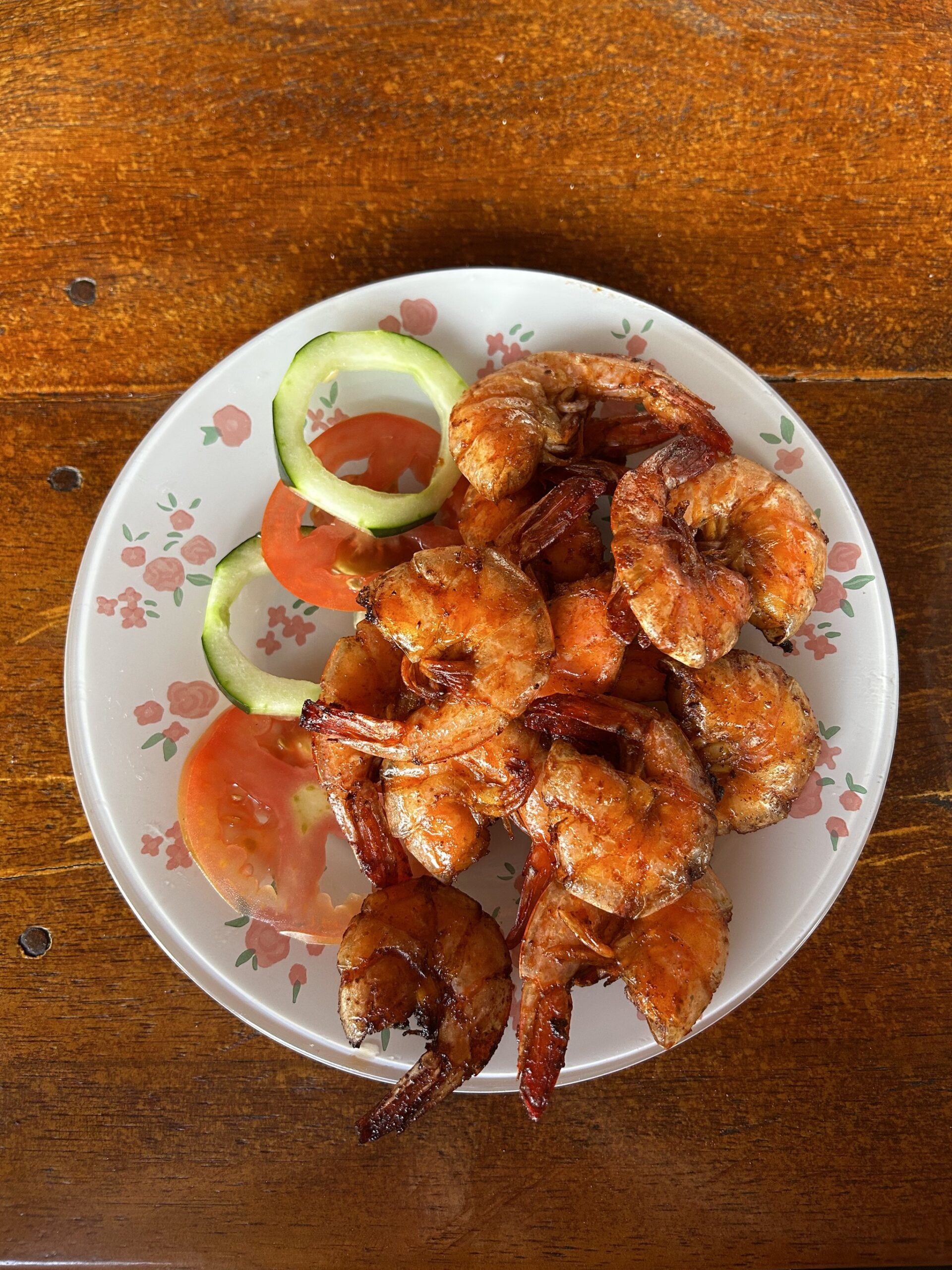 Fresh, fire-kissed shrimp — a taste of Maranhão on your plate. A plate of golden-red seasoned grilled shrimp garnished with tomato and cucumber slices, served on a floral-rimmed plate at a local restaurant near Lençóis Maranhenses Santo Amaro