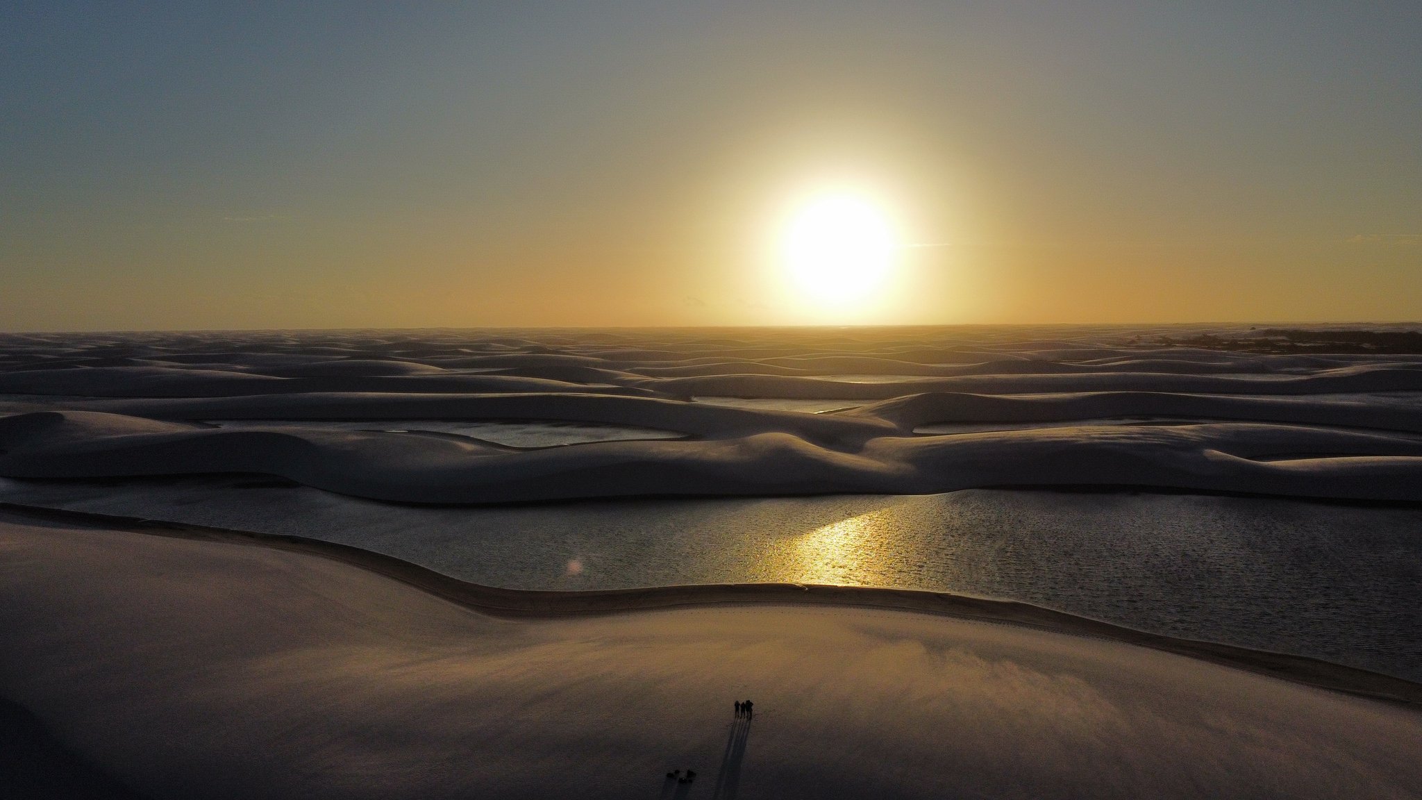 Vista aérea de drone das dunas de areia branca e lagoas espelhadas dos Lençóis Maranhenses banhadas pela luz dourada do pôr do sol, com duas silhuetas minúsculas projetando longas sombras
