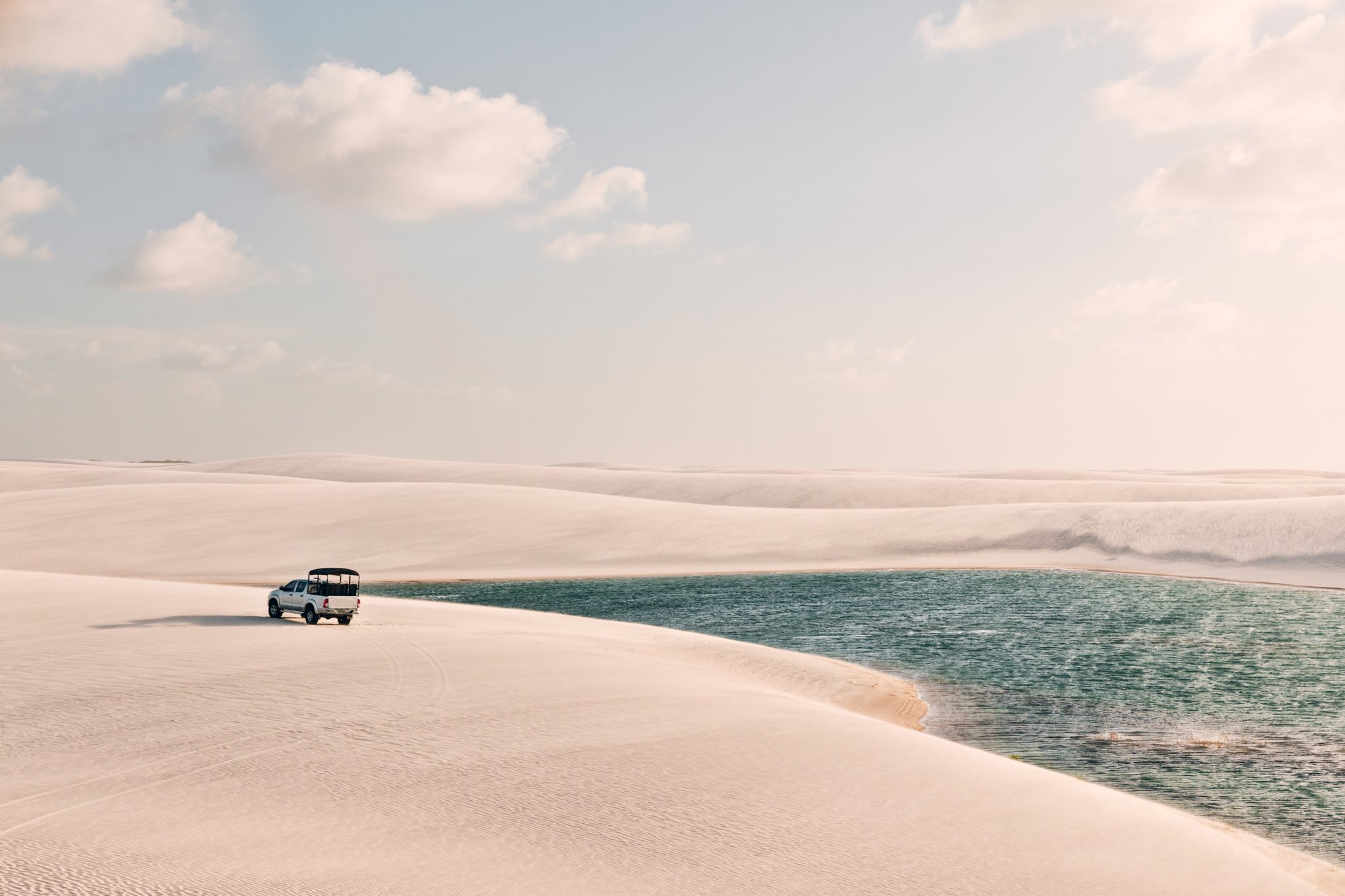 A white 4x4 pickup truck drives along the edge of a turquoise rainwater lagoon surrounded by vast white sand dunes in Lençóis Maranhenses, Brazil