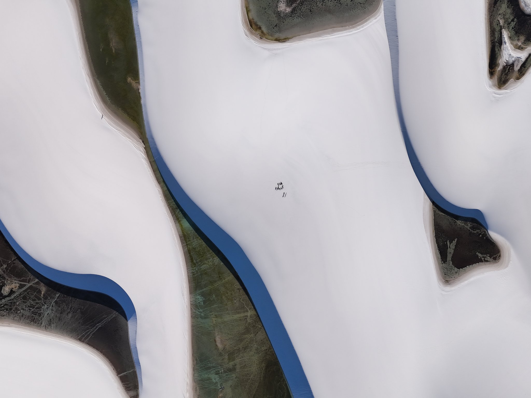 Vista aérea diretamente de cima do terreno dos passeios em Lençóis Maranhenses mostrando vastas dunas de areia branca separadas por canais sinuosos de água escura e lagoas parcialmente cobertas, com pequenas figuras humanas visíveis ao centro