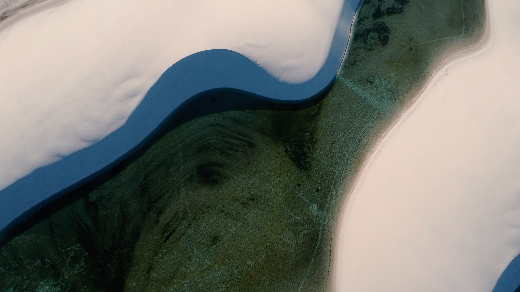 Aerial view of a deep blue lagoon cutting through white sand dunes in Lençóis Maranhenses, with a green seagrass-covered sandbar visible beneath the clear water