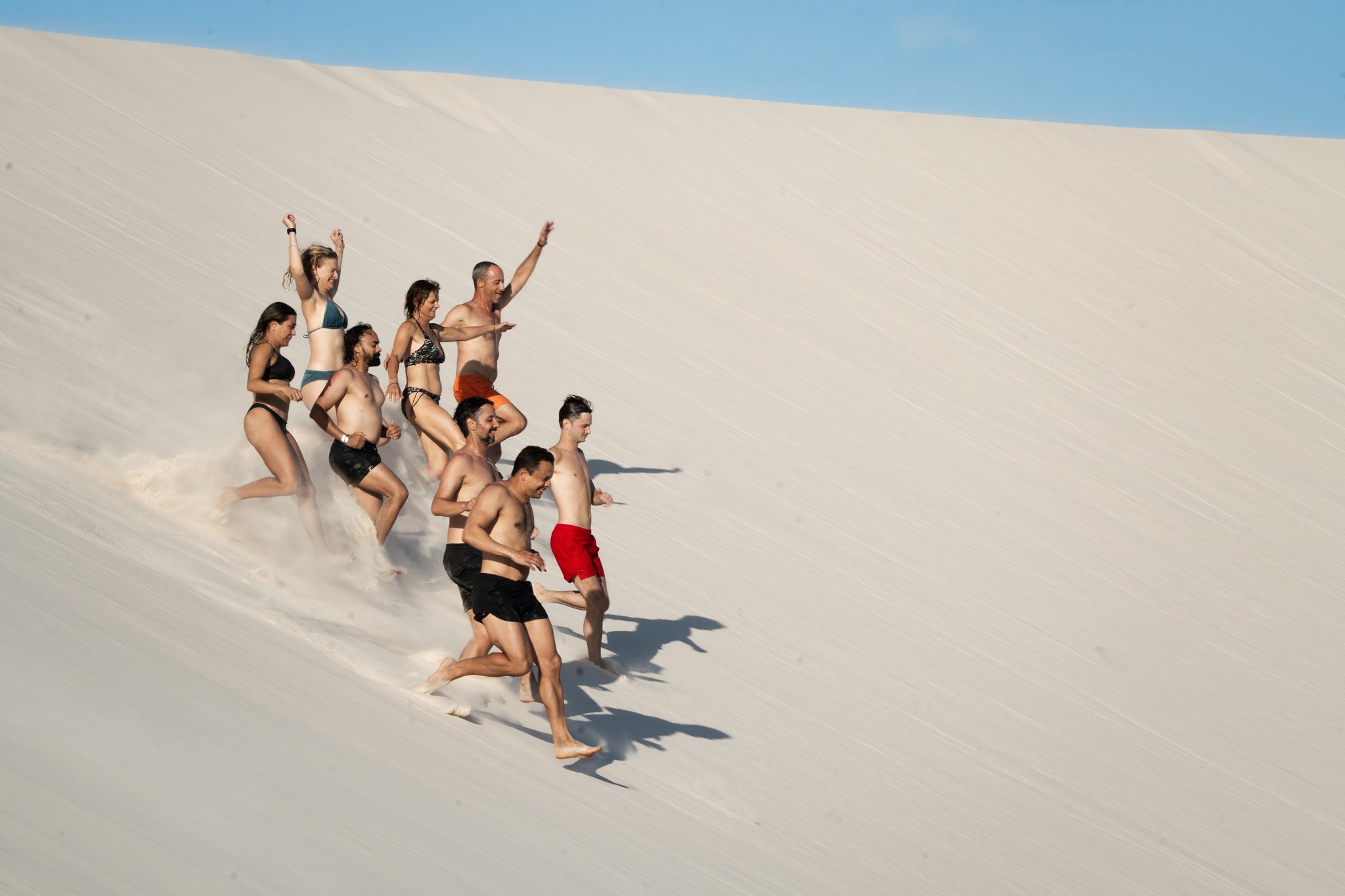 A group of eight travelers in swimwear laughing and sprinting barefoot down the steep white sand dunes of Lençóis Maranhenses tours in Brazil
