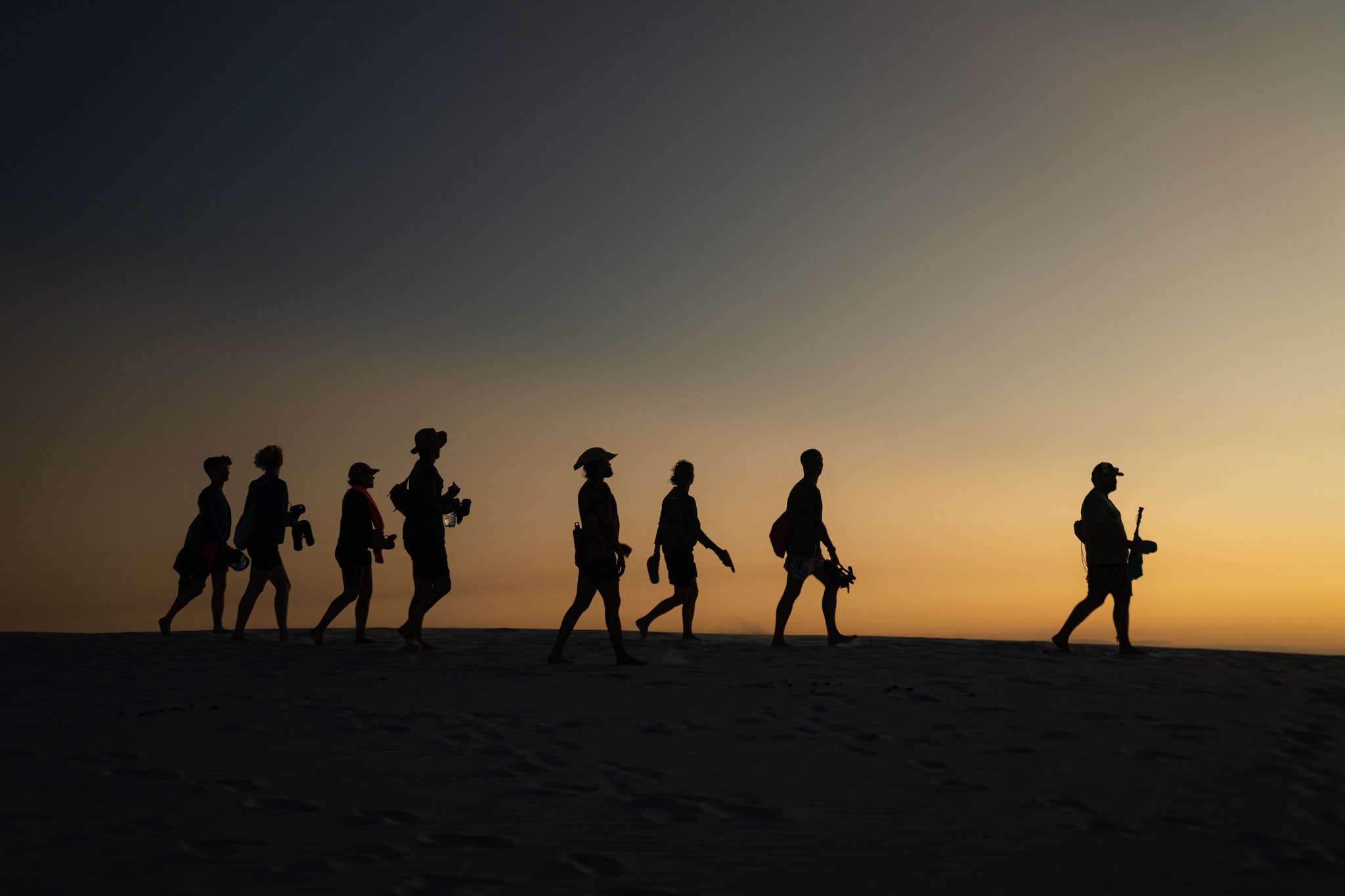 Eight travelers silhouetted against a vivid orange sunset sky as they walk single-file across white sand dunes on a Lençóis Maranhenses tour