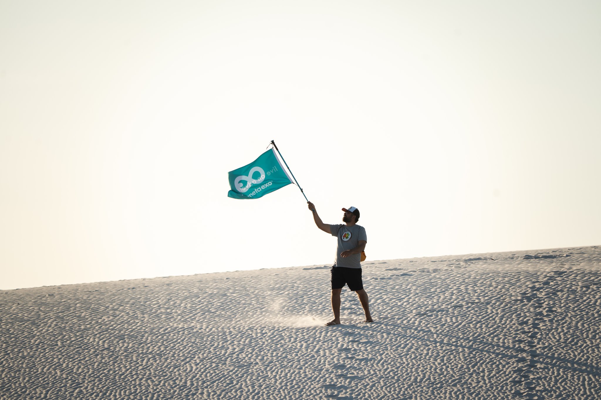 A tour guide waves a teal PlanetaEXO flag on top of a white sand dune in Lençóis Maranhenses, Brazil