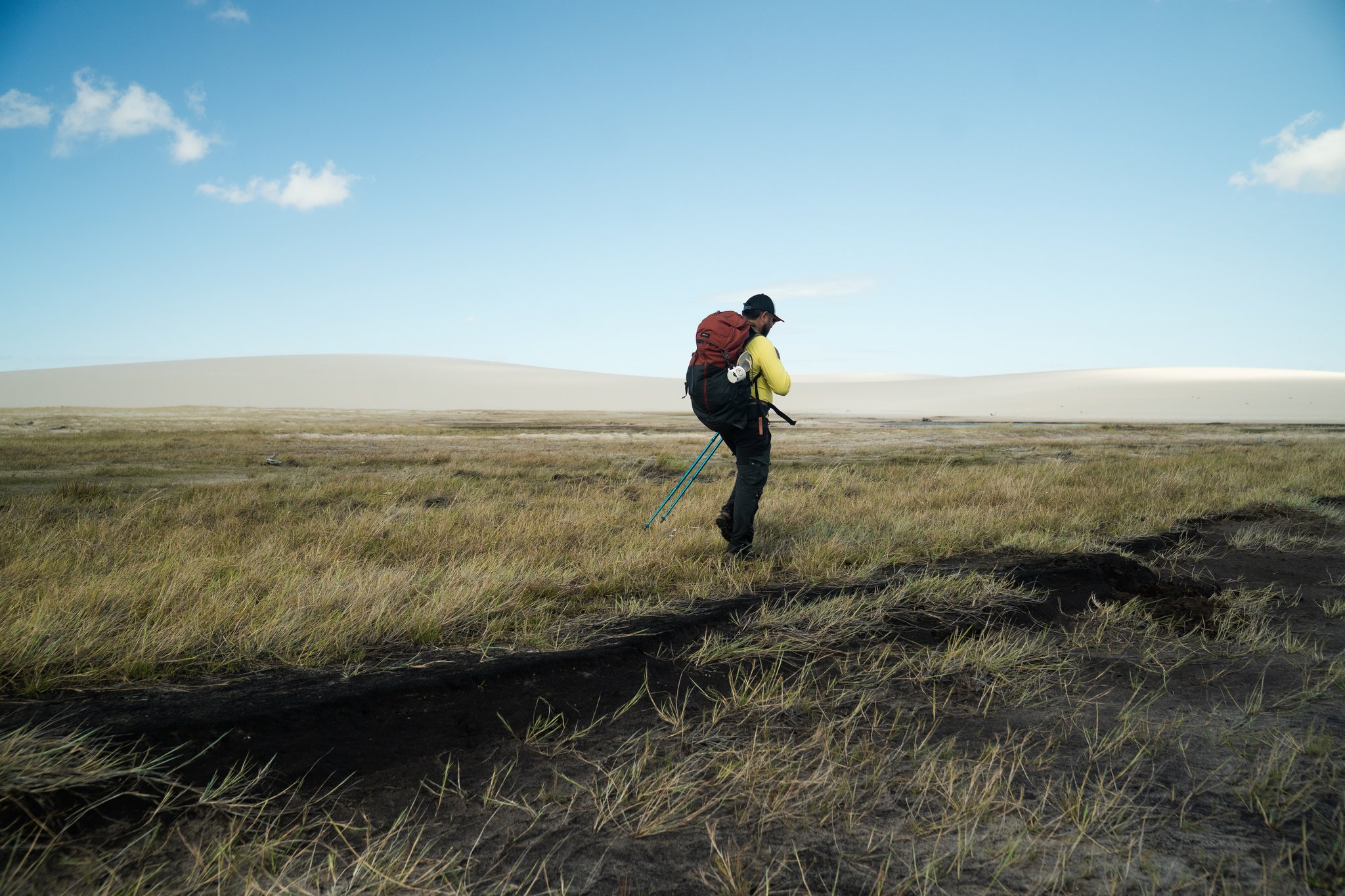 A hiker with a red backpack and trekking poles crosses a windswept grassland flat on a lençóis maranhenses tour, with pale sand dunes rising along the horizon under a blue sky