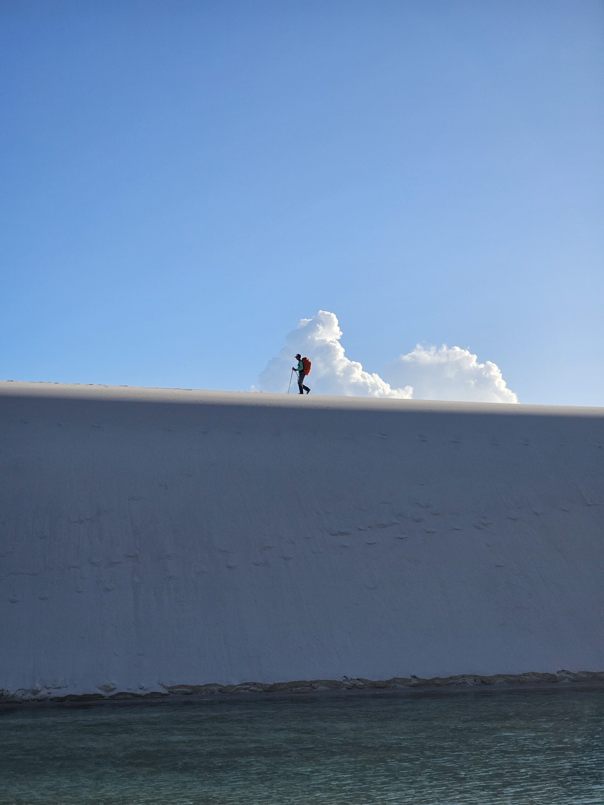 Um caminhante solitário com mochila vermelha e bastão de trekking percorre o cume afiado de uma enorme duna de areia branca em um passeio pelos Lençóis Maranhenses, com água turquesa da lagoa abaixo e nuvens cúmulos no horizonte