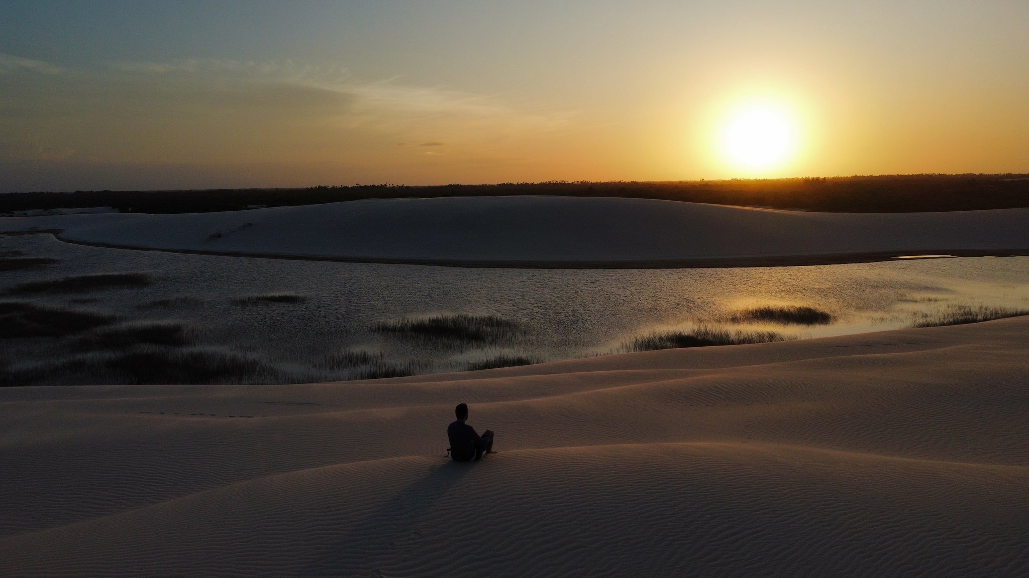 A lone traveler sits silhouetted on a white sand dune overlooking a still lagoon at golden hour during a lençóis maranhenses tour