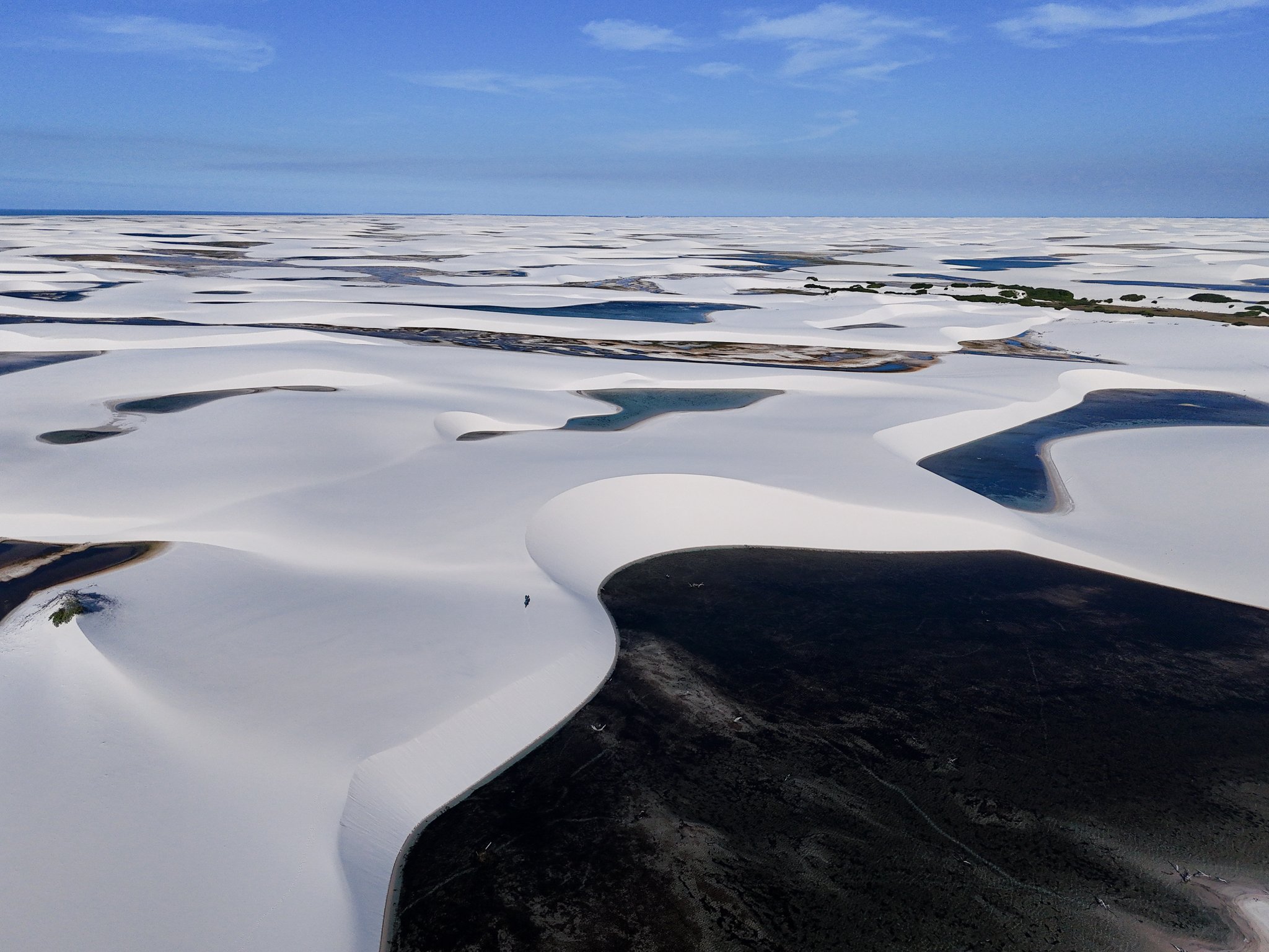 Sculpted by wind and rain: an ocean of white dunes and hidden lagoons, reaching as far as the eye can see. Aerial view of the Lençóis Maranhenses Trek from Atins, showing vast white sand dunes stretching to the horizon with dark and blue freshwater lagoons nestled between them under a clear blue sky