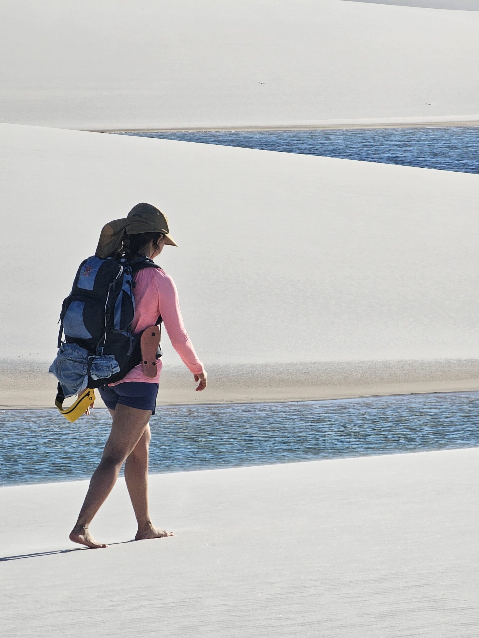Barefoot between dune and lagoon — the only way to travel. A barefoot woman with a backpack walks along the edge of a blue lagoon framed by sweeping white sand dunes on the Lençóis Maranhenses trek from Atins