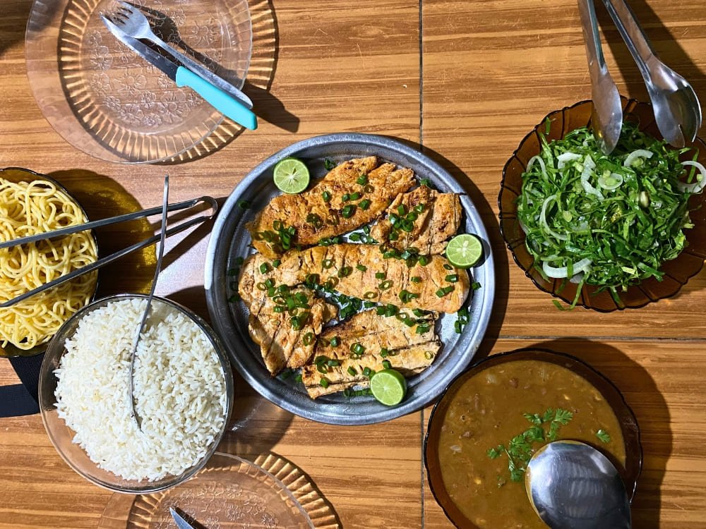 Fresh, local flavours cooked by hand — fuel for the dunes ahead. A spread of home-cooked food on a wooden table during the Lencois Maranhenses Trek Atins experience, featuring grilled fish fillets garnished with lime and spring onion, white rice, a green herb salad, bean stew, and noodles