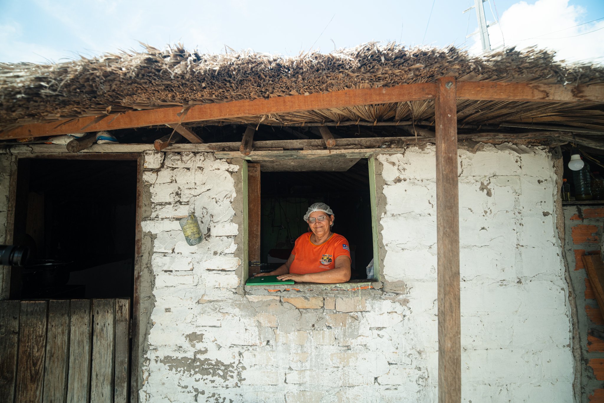 A smiling woman wearing an orange shirt and hairnet leans out of the window of a rustic whitewashed brick house with a thatched palm roof, part of the Lençóis Maranhenses trek from Atins homestay experience