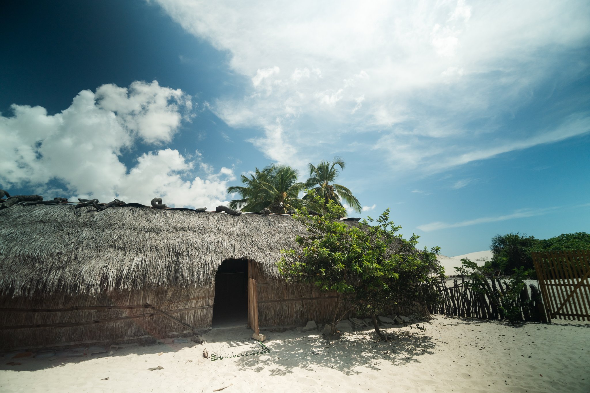 Simple, sun-bleached walls and a palm canopy — life inside the dunes. A rustic thatched-roof dwelling on white sand, surrounded by tropical shrubs and palm trees under a bright blue sky on the Lençóis Maranhenses trek from Atins