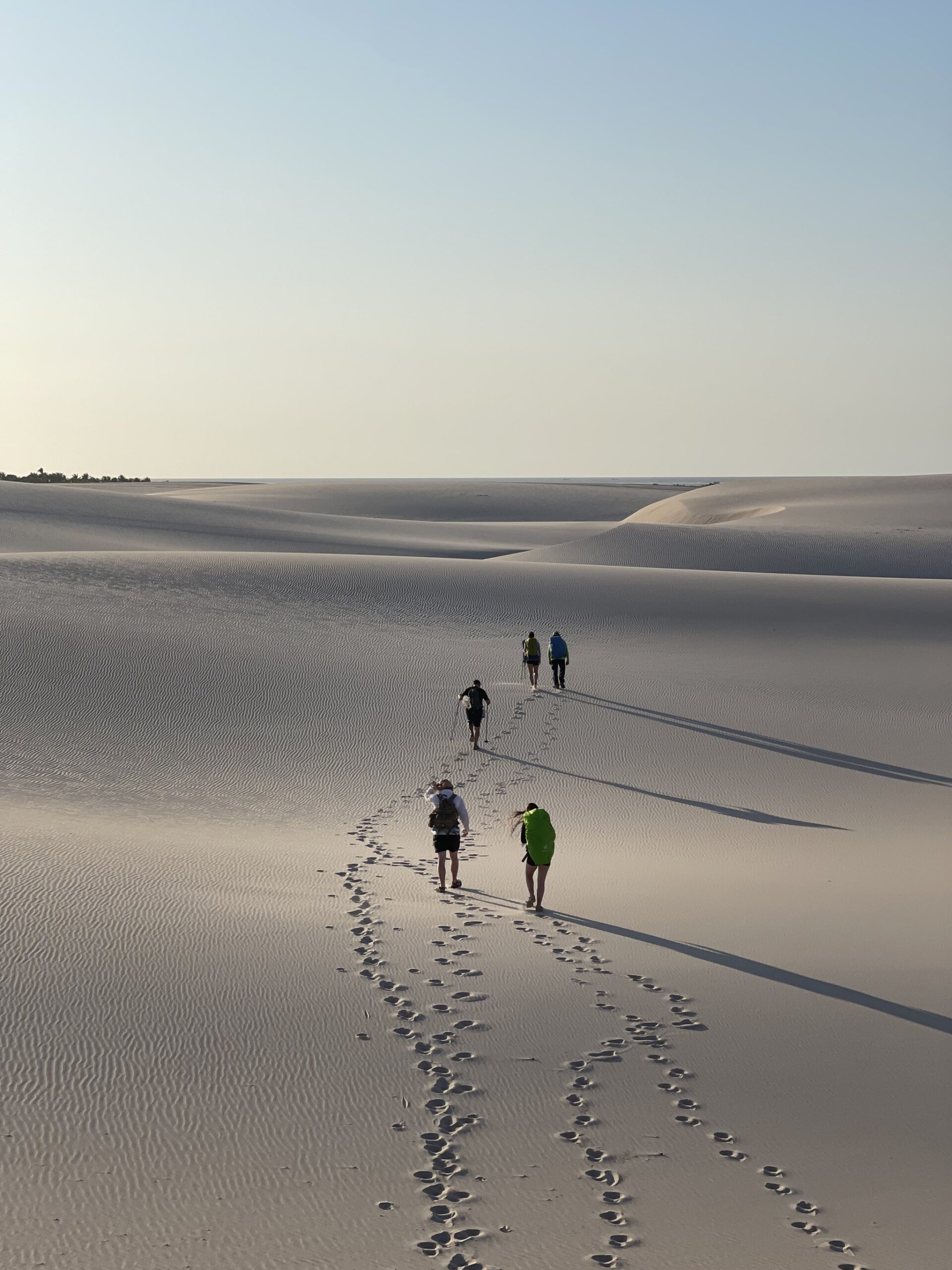 Four packs, one horizon — the dunes stretch endlessly ahead. Four trekkers with backpacks walking single-file across vast rippled white sand dunes on a lençóis maranhenses trek, their footprints trailing behind them toward the horizon