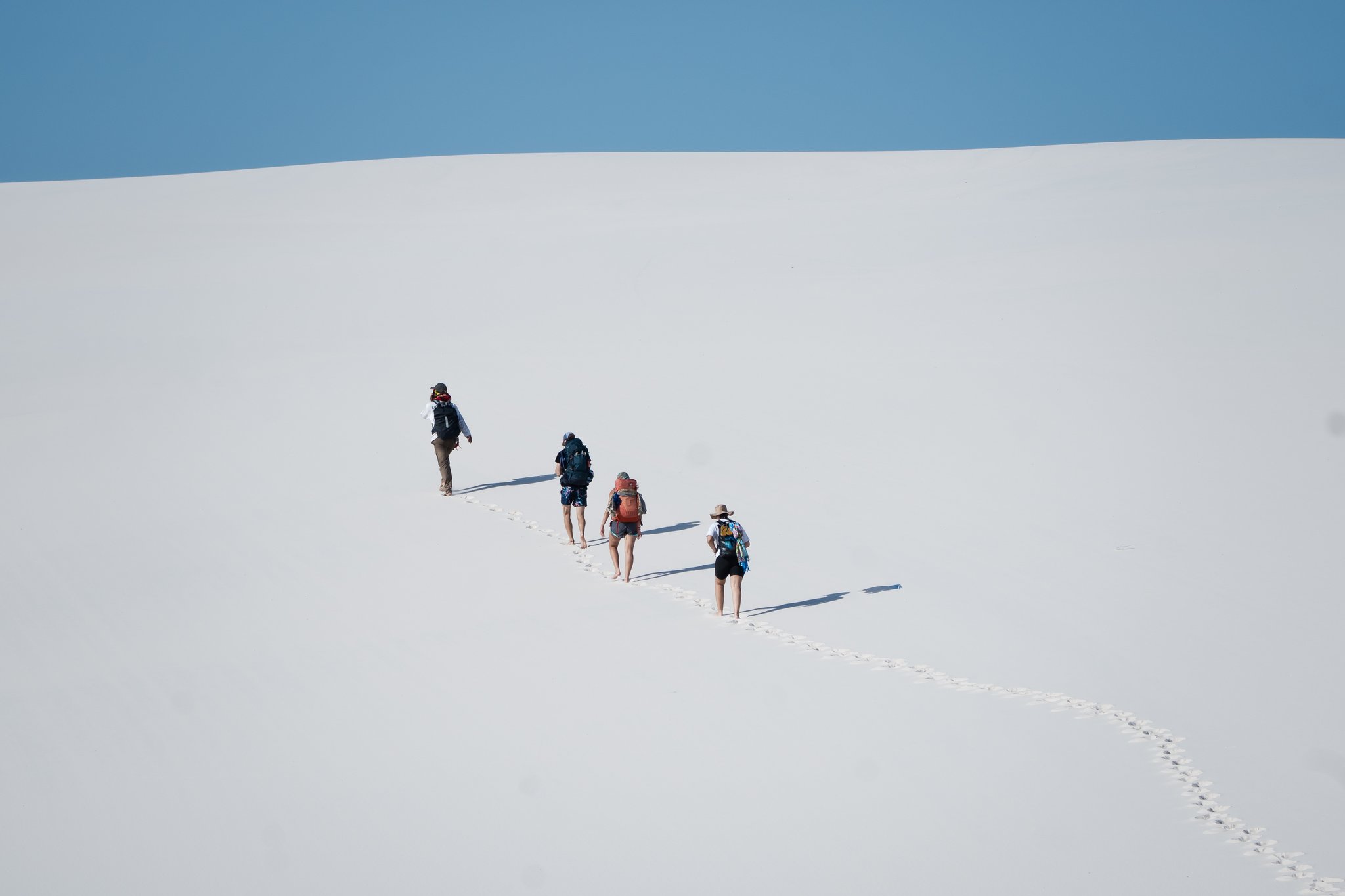 Footprints in the white, stretching toward the ridge. Four backpackers climbing a vast white sand dune in single file during a lencois maranhenses trek, their footprints trailing behind them beneath a clear blue sky