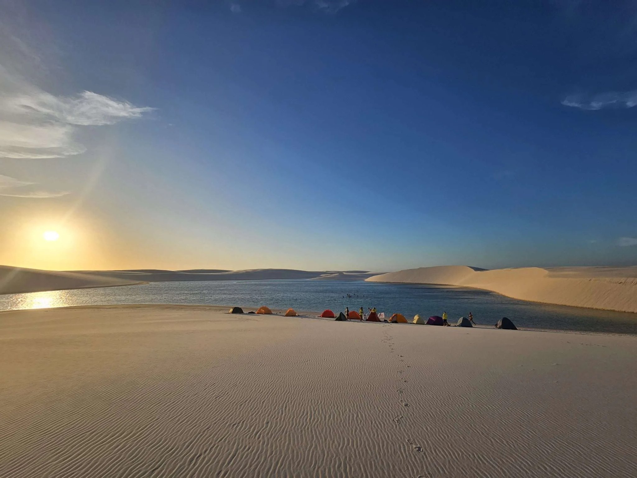 Colorful tents lined along a freshwater lagoon at sunset during a lençóis maranhenses trek, with rippled white sand dunes stretching to the horizon under a deep blue sky