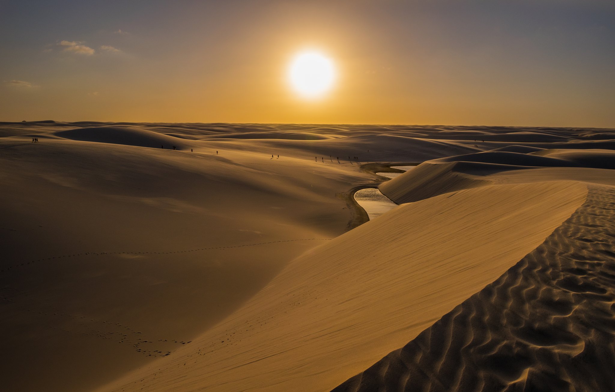 Sun melting into the dunes, a lagoon catching the last light — the lençóis maranhenses trek at its most elemental. Photo: NidoHuebl Golden sunset blazing over an endless sea of white sand dunes in Lençóis Maranhenses, with a narrow freshwater lagoon winding between the slopes and tiny silhouettes of trekkers visible on the crests in the distance