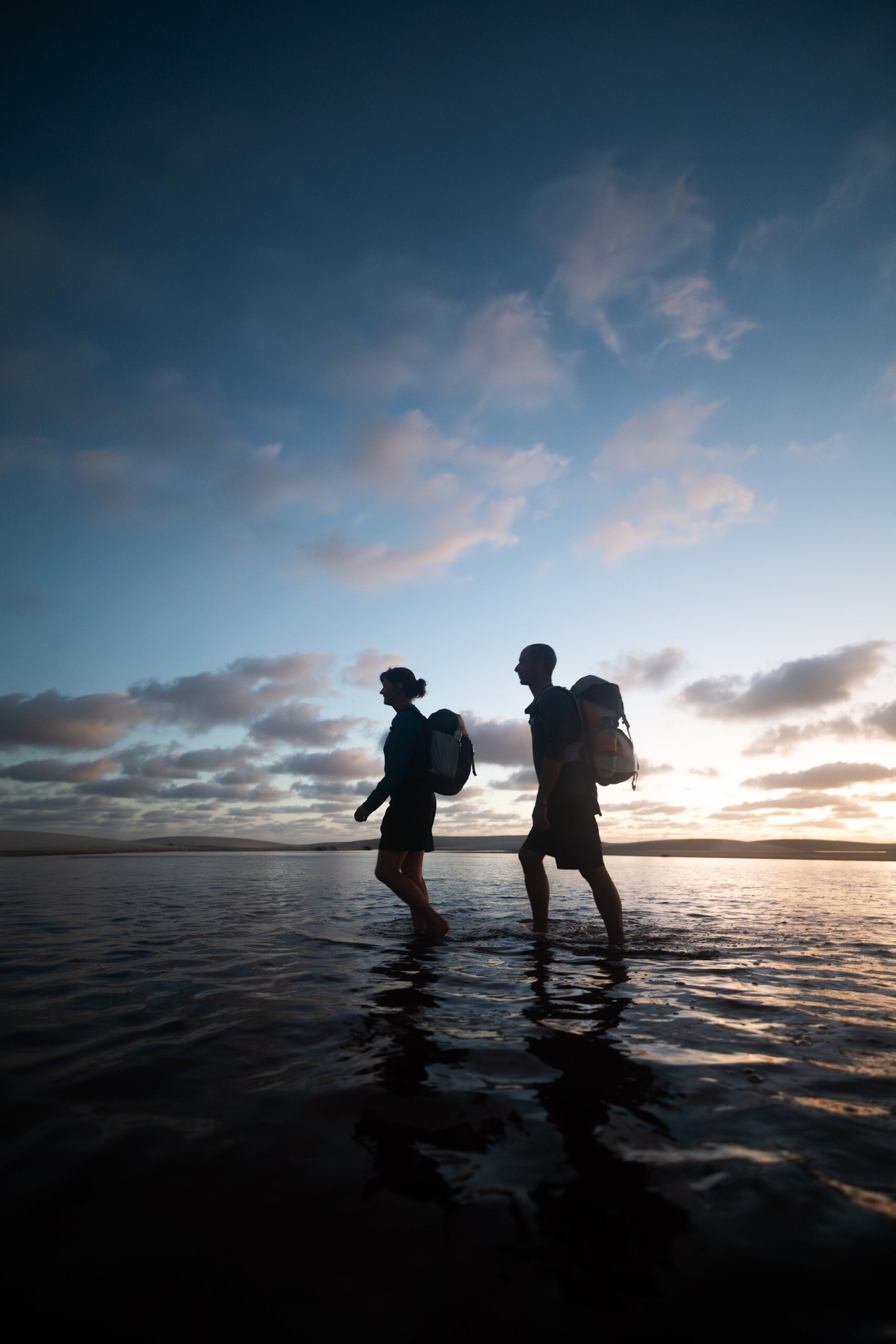 Dusk water underfoot, the far shore still hours away. Two trekkers carrying backpacks wade barefoot through shallow water at dusk on the lencois maranhenses trek, silhouetted against a wide twilight sky reflected on the still surface
