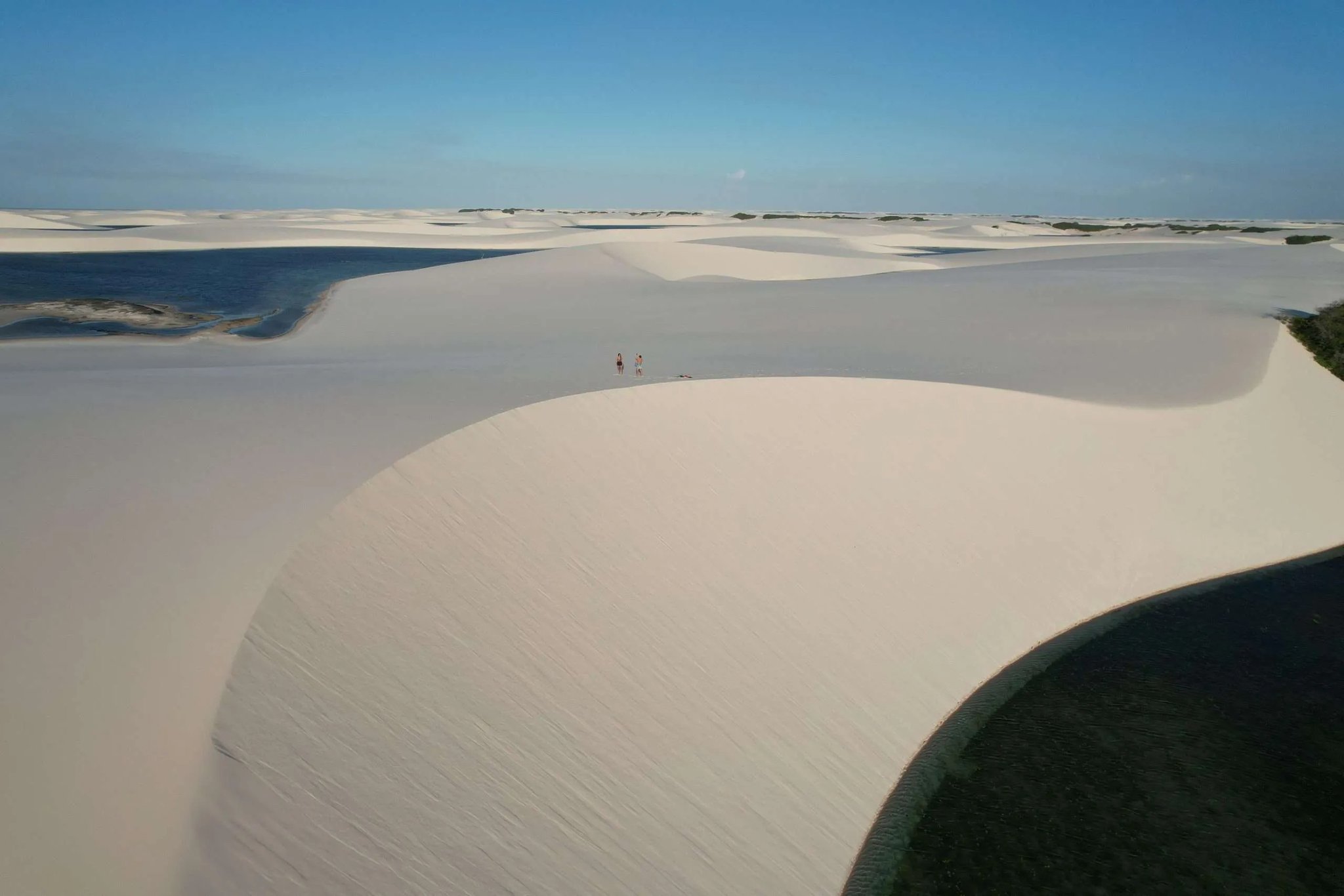 Humans reduced to specks — the dunefield sets the scale. Two tiny figures stand atop a sweeping white dune crest during a lençóis maranhenses trek, surrounded by a mosaic of glassy blue lagoons and rolling sand stretching to the horizon