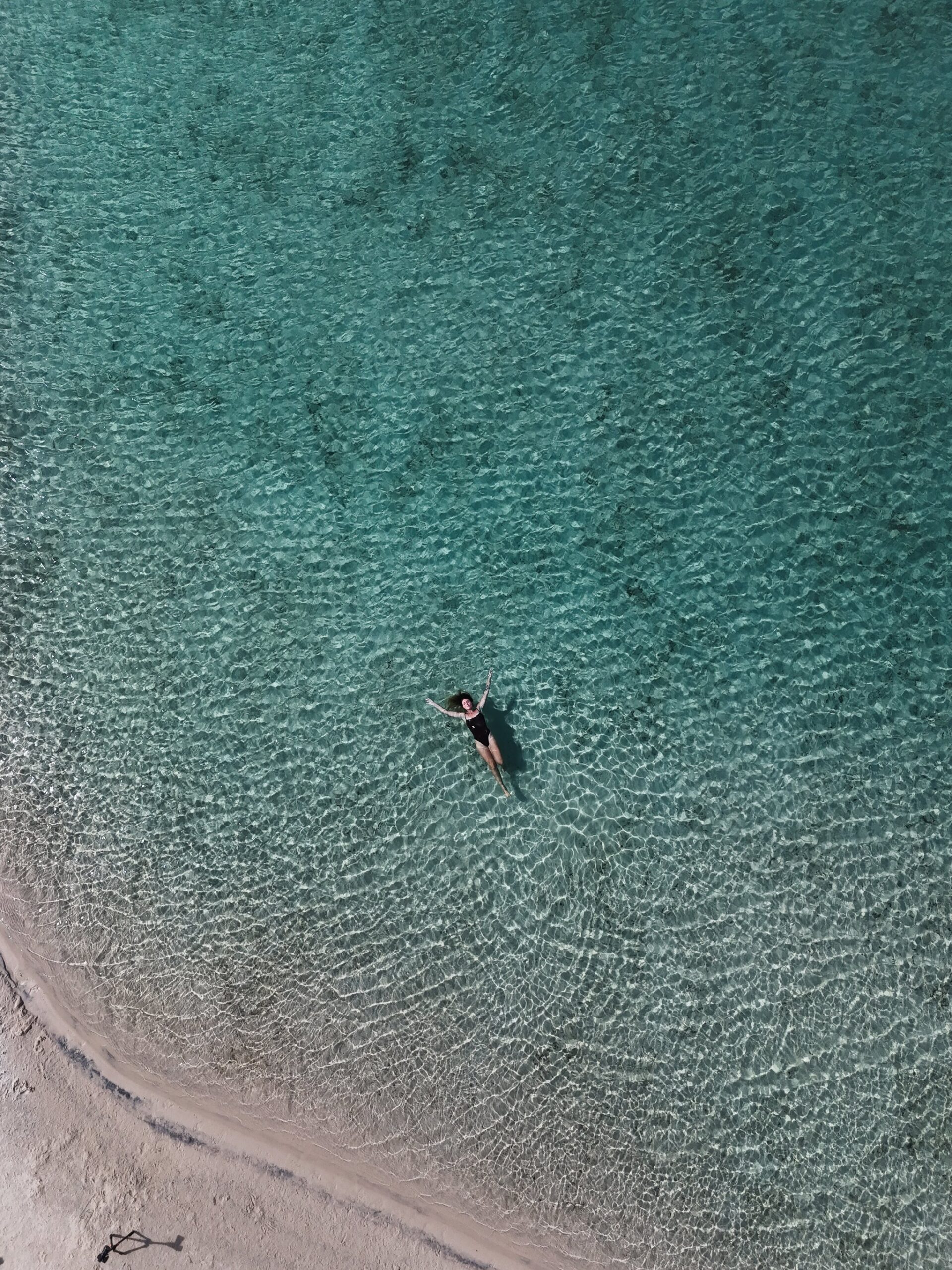 Arms wide open, adrift in pure turquoise. Aerial view of a woman floating on her back in shallow turquoise water beside a white sand shore during a Lençóis Maranhenses trekking experience