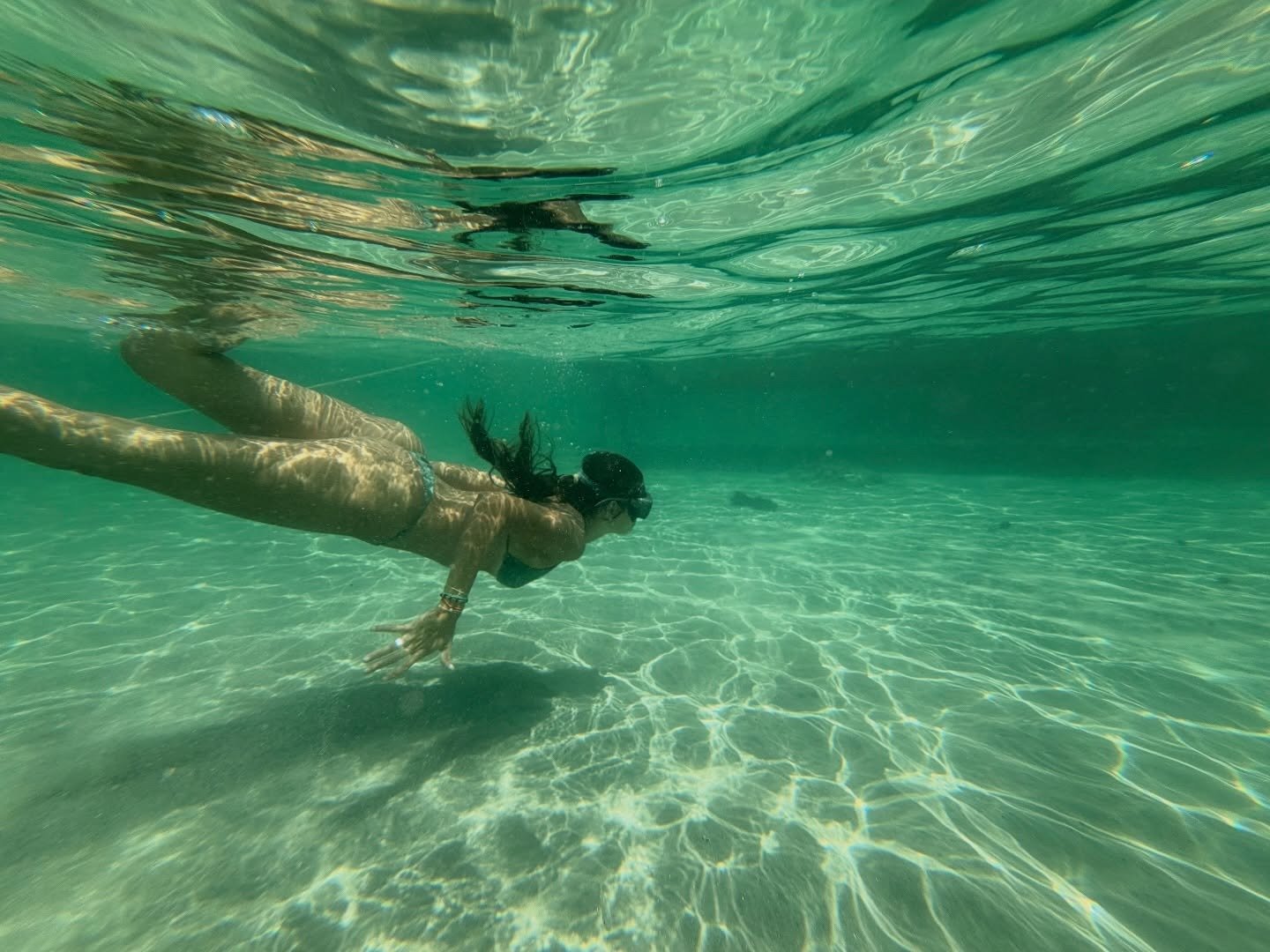 A woman free-dives through crystal-clear turquoise water above a white sandy bottom during a Lençóis Maranhenses trekking lagoon stop
